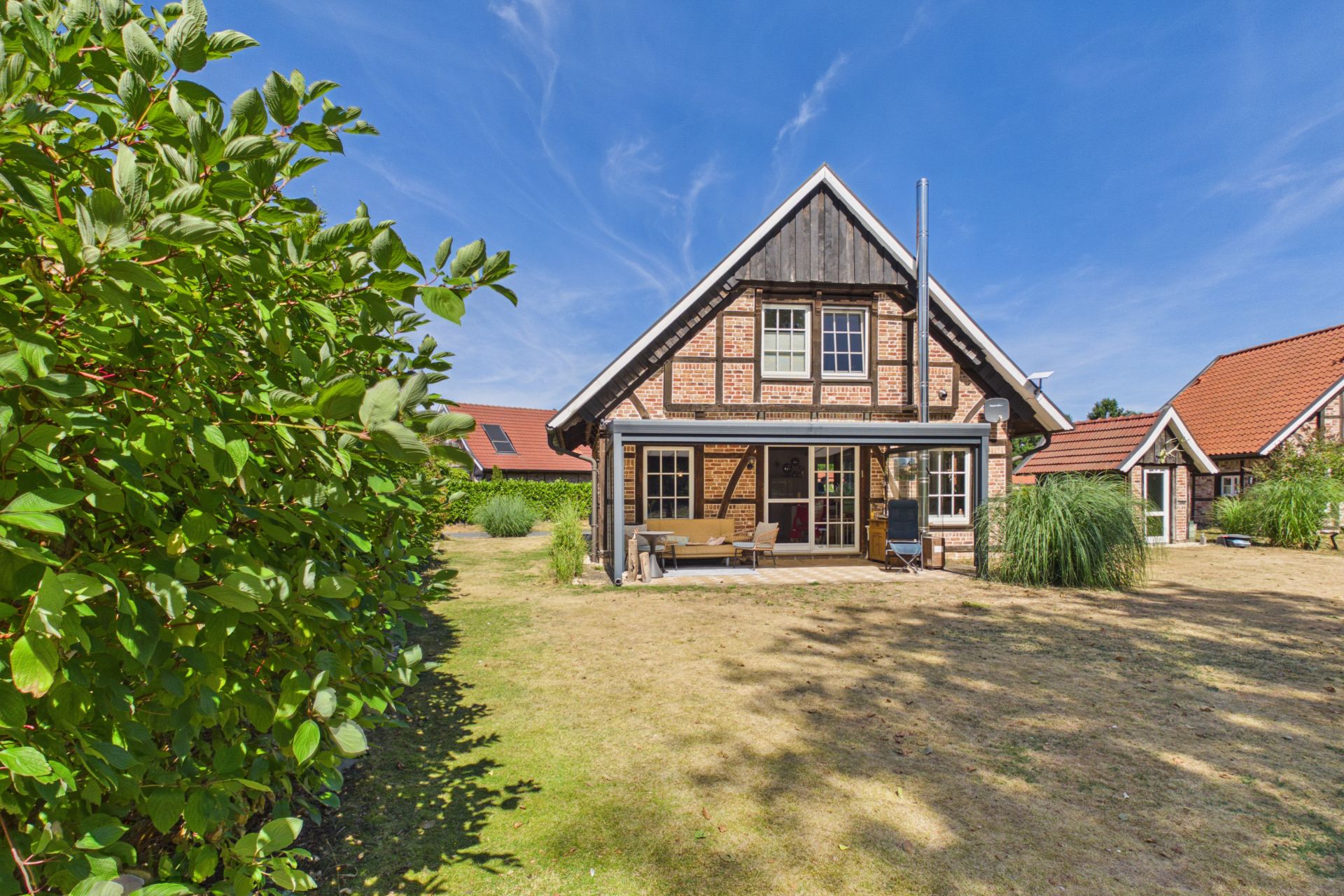 Fachwerkhaus mit Terrasse und Garten, blauer Himmel, Sträucher und Rasen im Vordergrund.