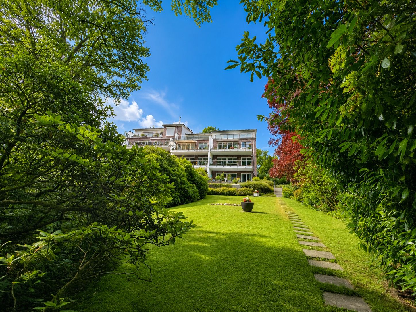Mehrfamilienhaus mit großen Balkonen, umgeben von grünem Garten und Bäumen unter blauem Himmel.