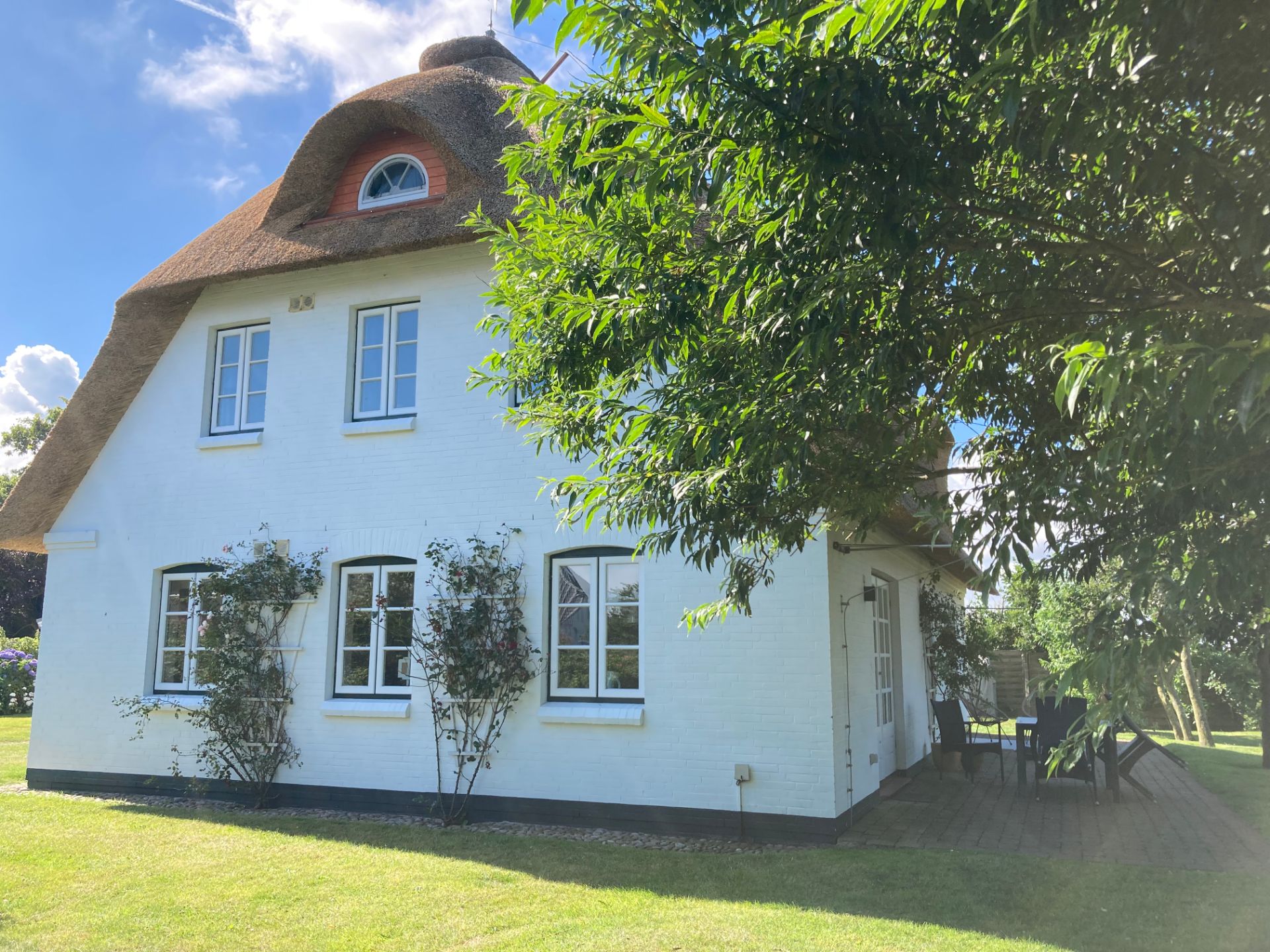 Weißes Haus mit Reetdach, mehreren Fenstern und einer Terrasse im Garten bei Sonnenschein.