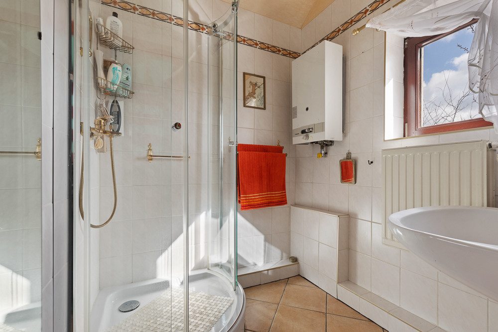 Kleines Badezimmer mit Eckdusche, Waschbecken, Heizung und Fenster mit Vorhang.