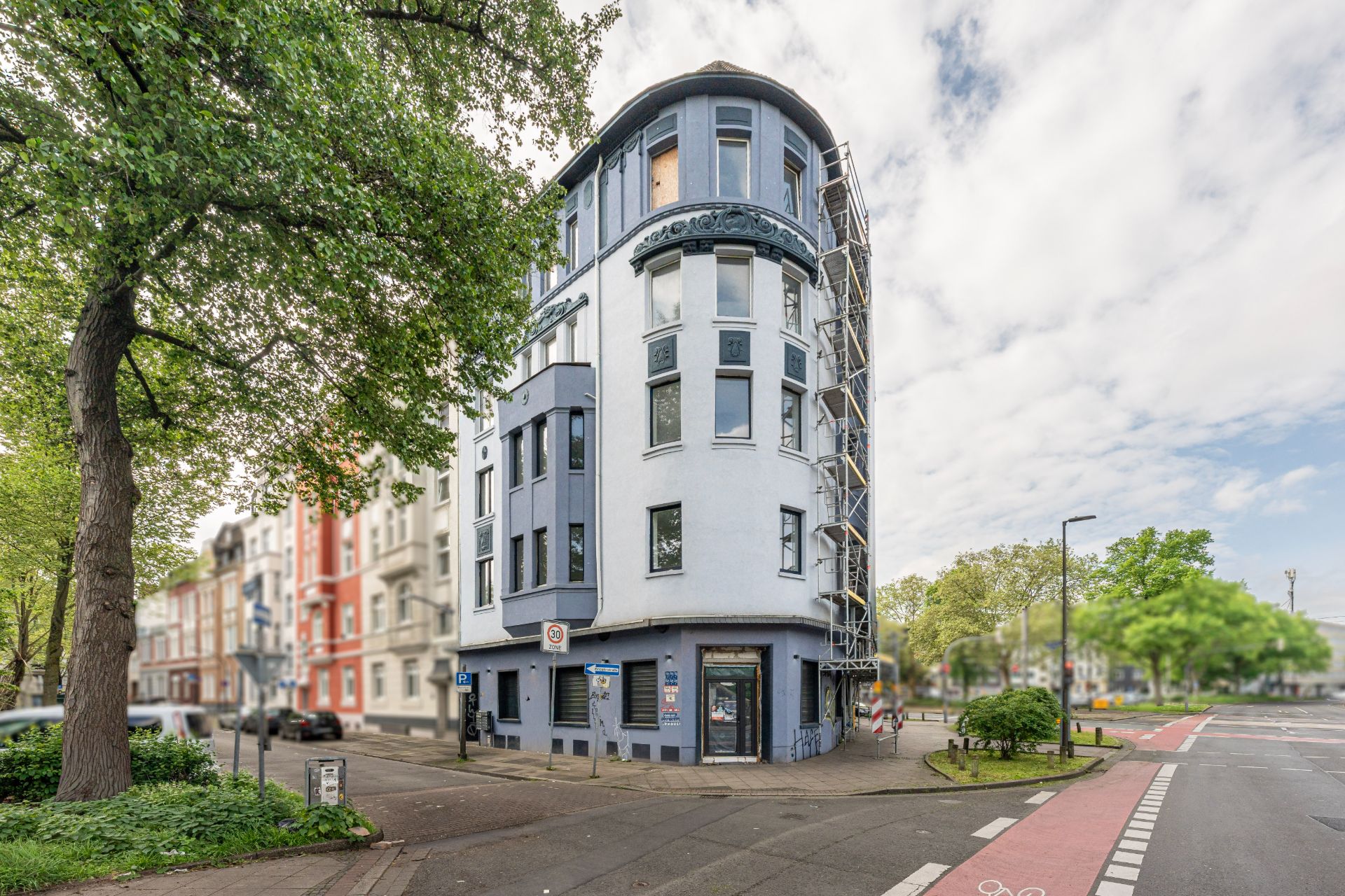 Eckhaus mit mehreren Stockwerken, blauer Fassade und vielen Fenstern an einer Straßenkreuzung.