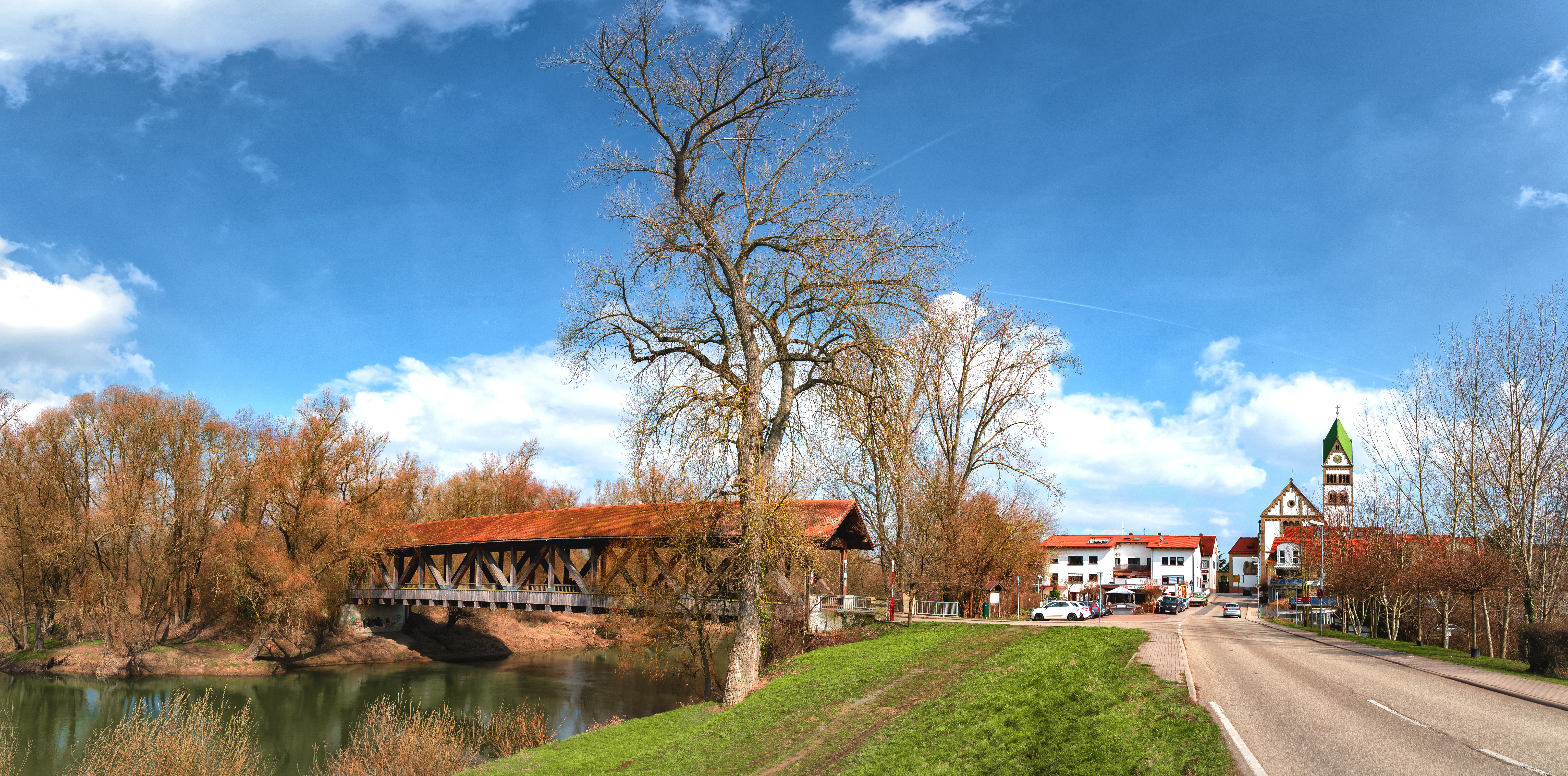 Panorama mit überdachter Holzbrücke, Fluss, Bäumen und Häusern unter blauem Himmel.