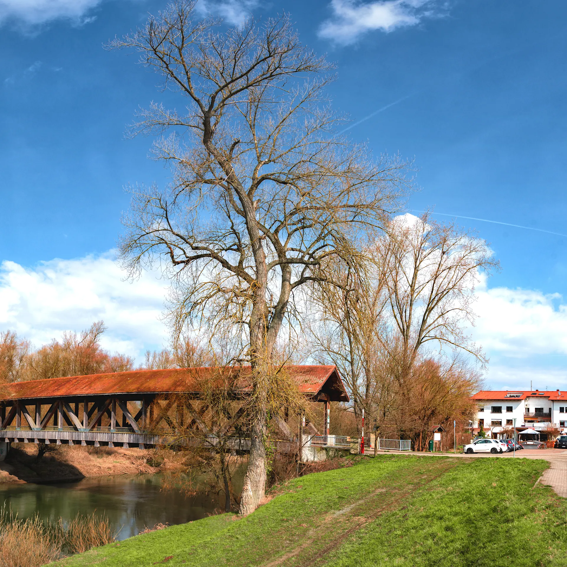 Panorama mit überdachter Holzbrücke, Fluss, Bäumen und Häusern unter blauem Himmel.