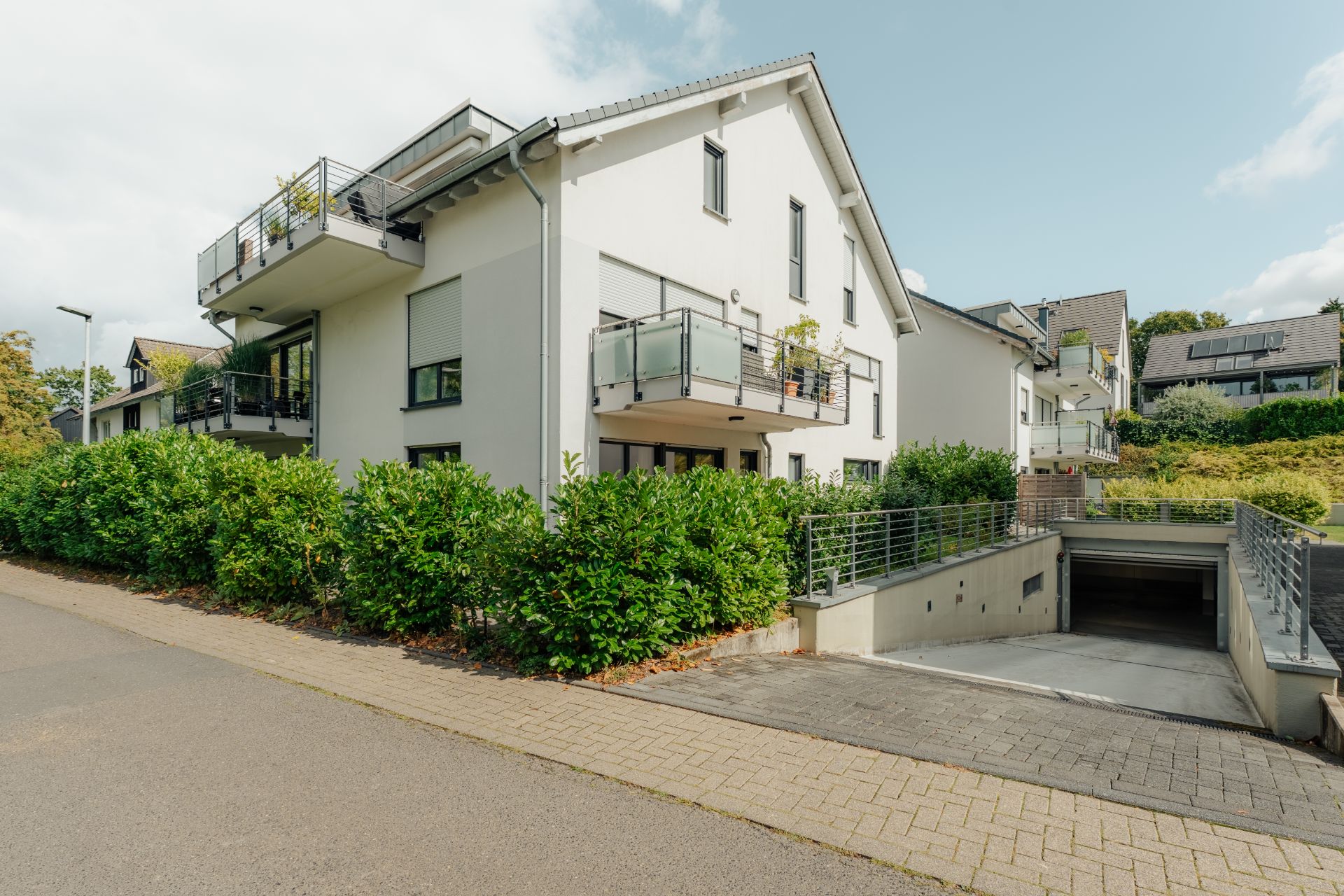 Modernes Wohnhaus mit Balkonen, Einfahrt zur Tiefgarage und grüner Hecke an der Straße.