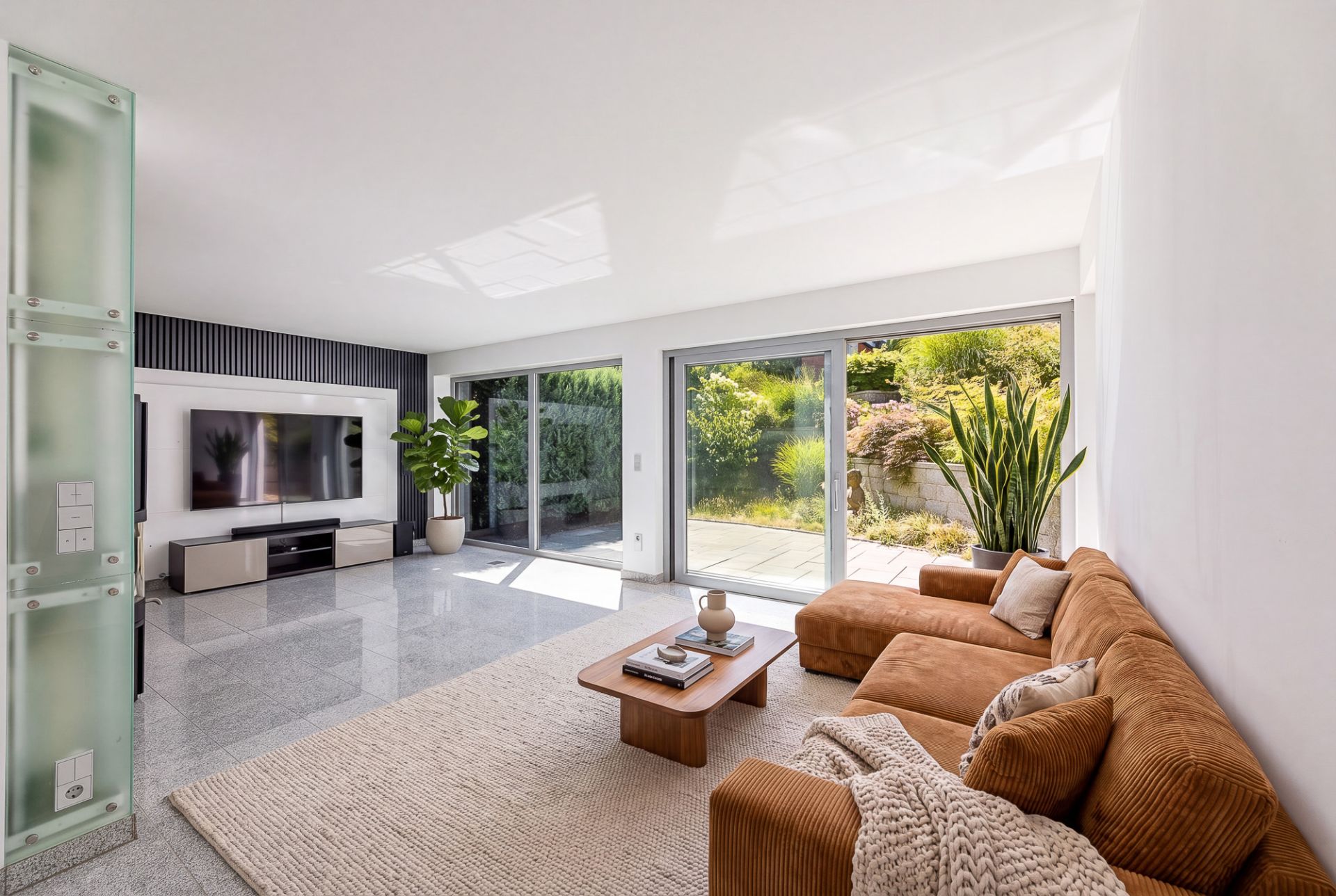 Helles Wohnzimmer mit braunem Sofa, Couchtisch, Teppich, TV und großen Fensterfronten zum Garten.