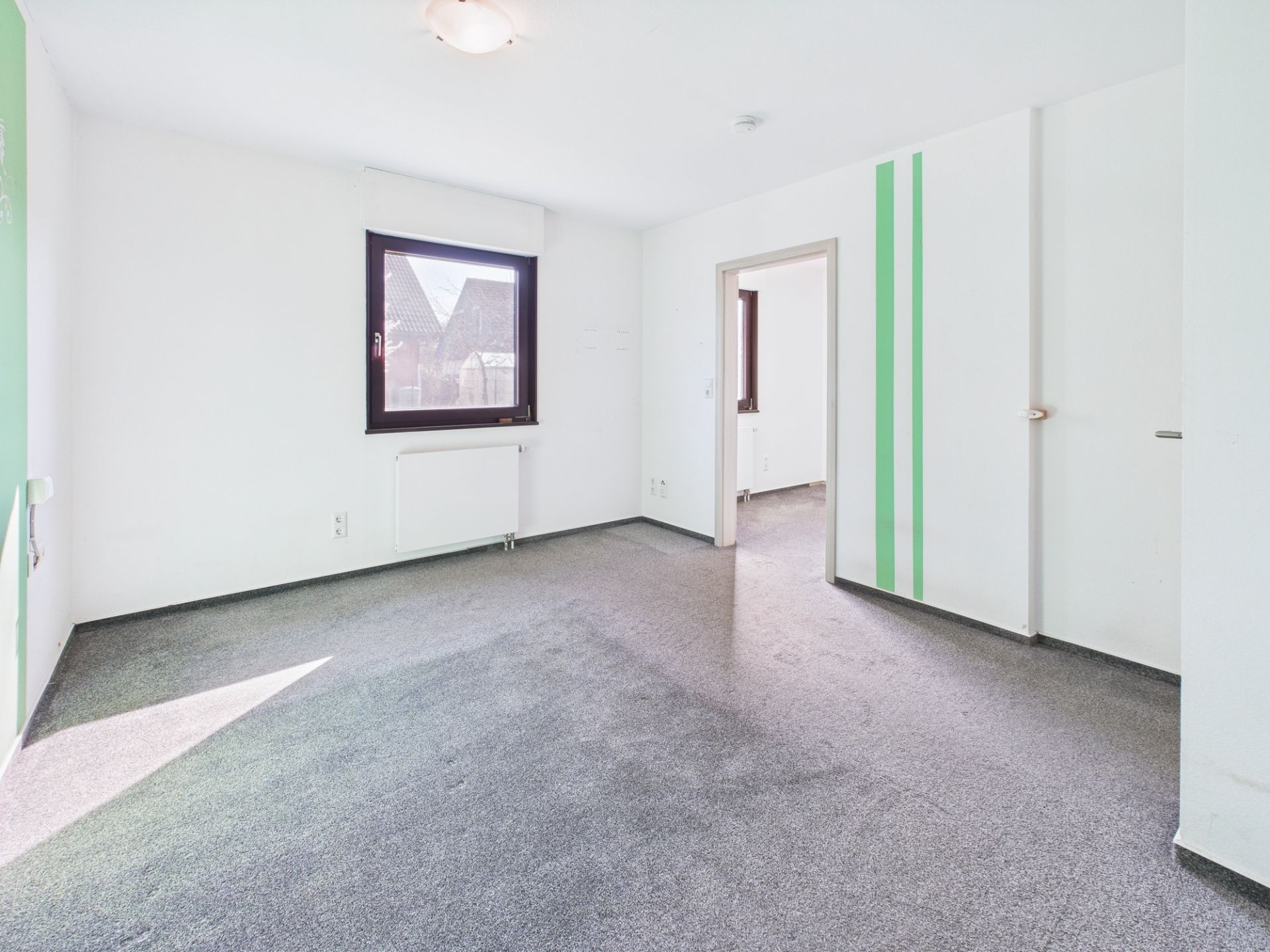 Heller, leerer Raum mit Teppichboden, Fenster und Tür zu weiterem Zimmer, weiße Wände mit grünen Streifen.