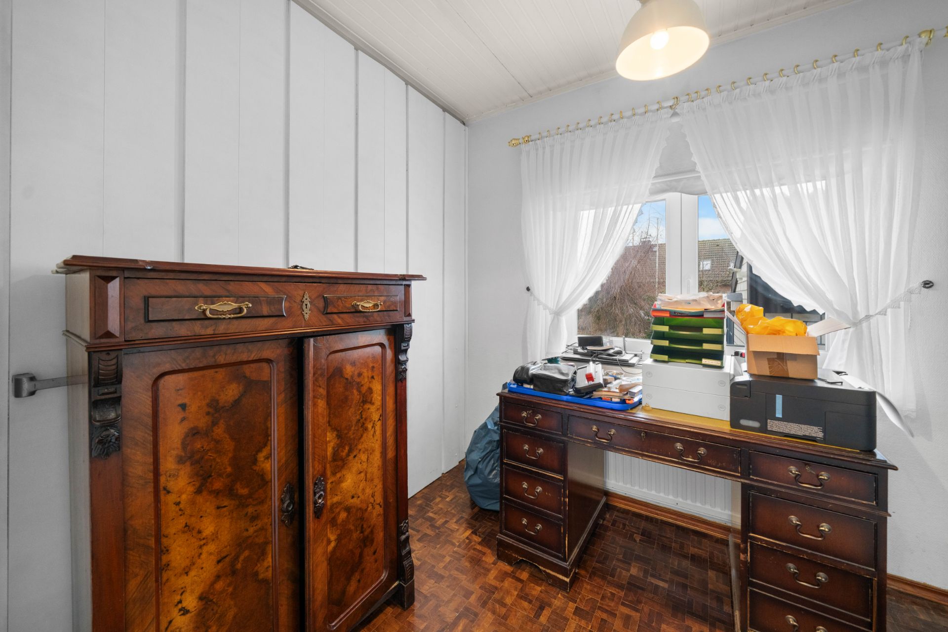 Kleines Zimmer mit altem Holzschrank, Schreibtisch und Fenstern mit weißen Vorhängen.