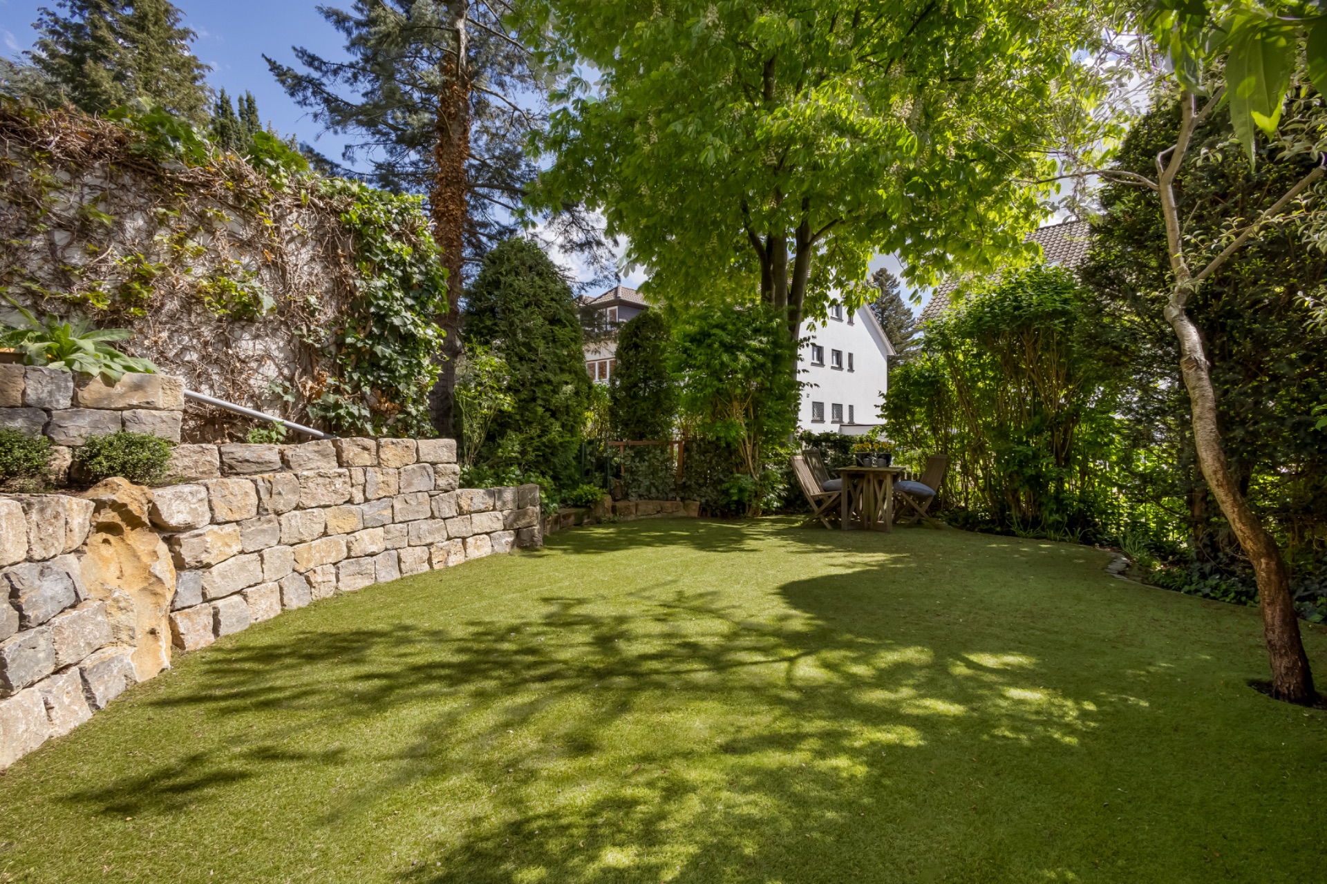 Grüner Garten mit Steintreppe, Rasenfläche und Sitzbereich unter Bäumen.