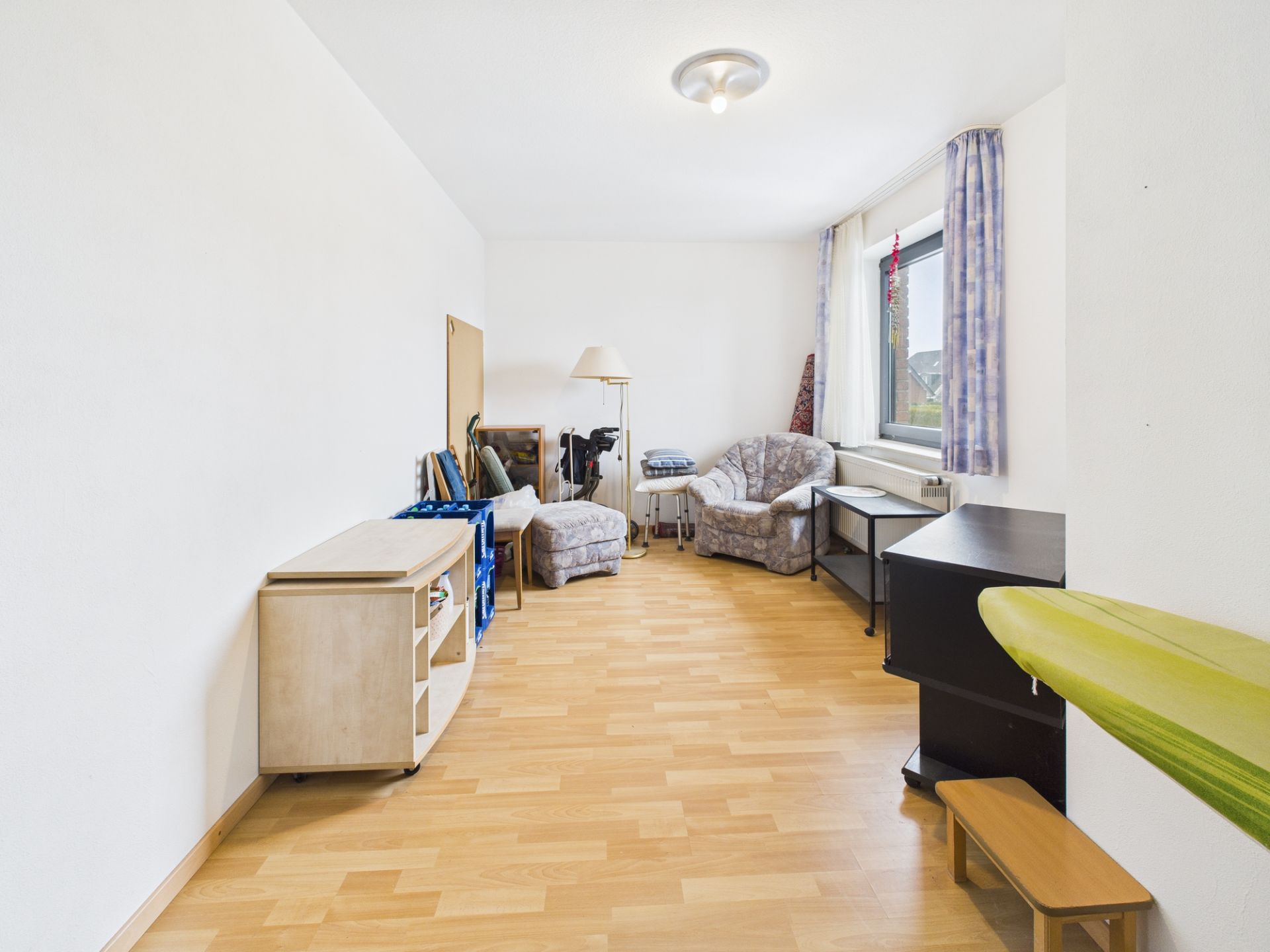 Helles Wohnzimmer mit Holzfußboden, Sesseln, Regal, Schreibtisch und großem Fenster mit Vorhängen.