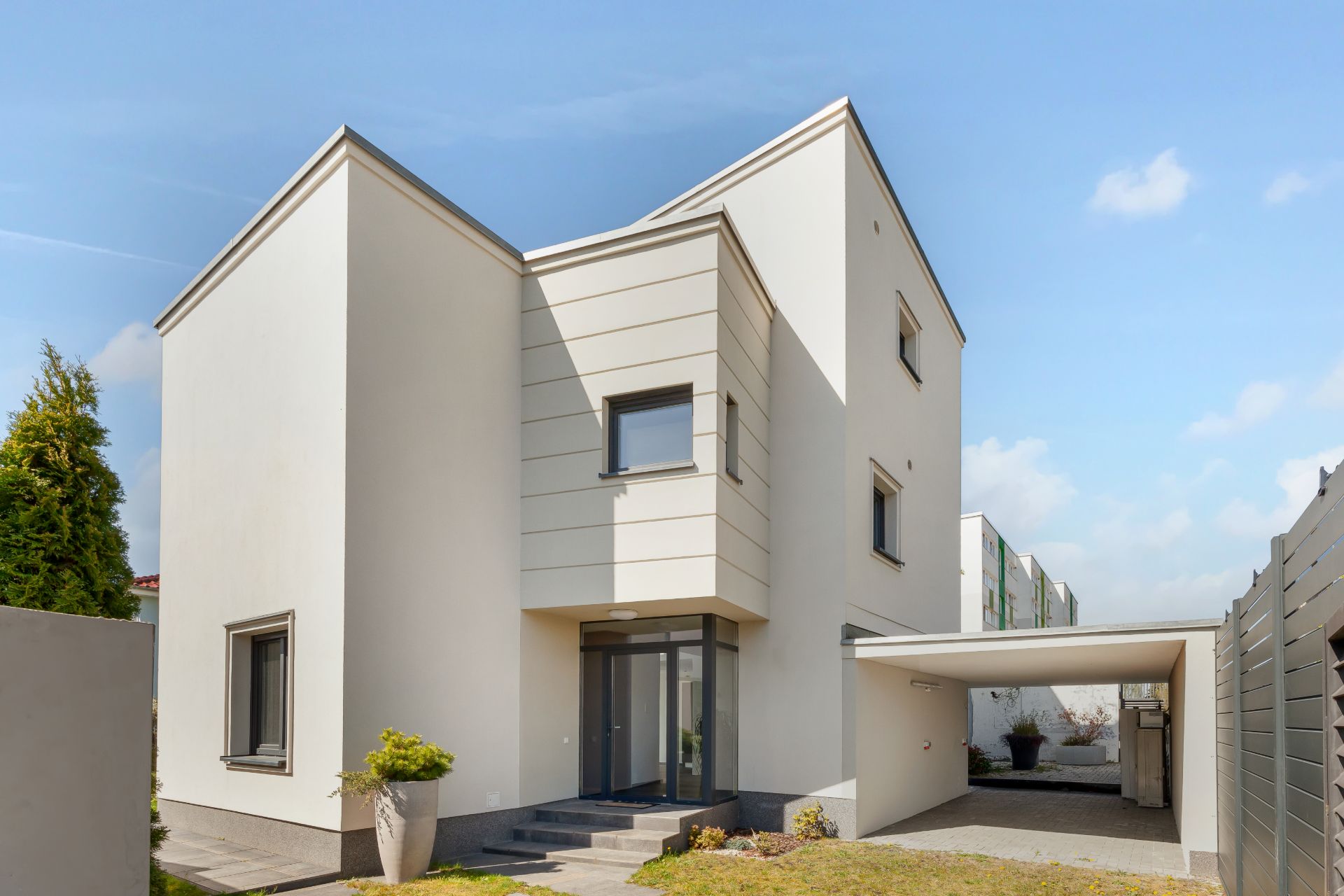 Modernes, weißes Wohnhaus mit Flachdach und Carport, umgeben von Garten und Einfahrt.