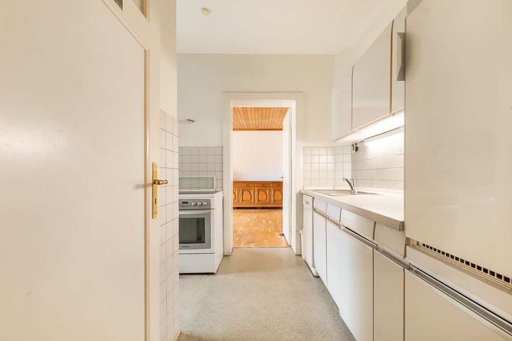 Helle schmale Küche mit weißen Schränken, Herd, Spüle und Blick in ein angrenzendes Zimmer.