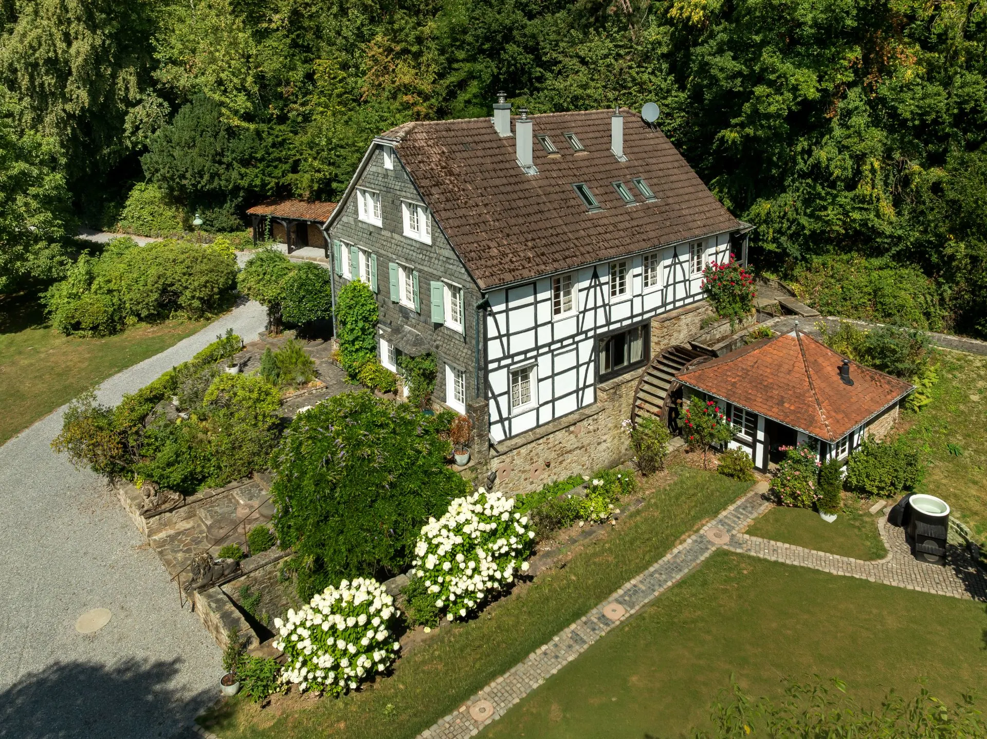 Fachwerkhaus mit Garten, Blumen und kleiner Nebengebäude, umgeben von Bäumen.
