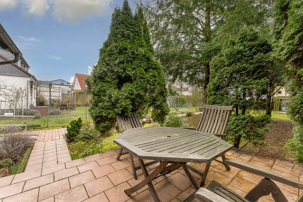 Gartenbereich mit Holztisch und Stühlen, umgeben von Bäumen und Gehweg aus Fliesen.