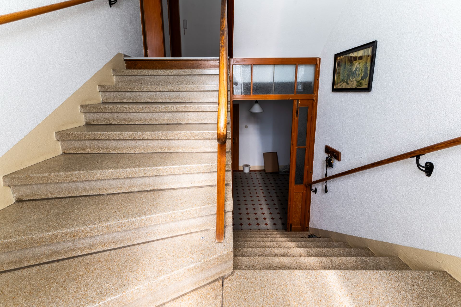 Treppenhaus mit Holzgeländer, weißer Wand, Bild an der Wand und Tür zu einem weiteren Raum.