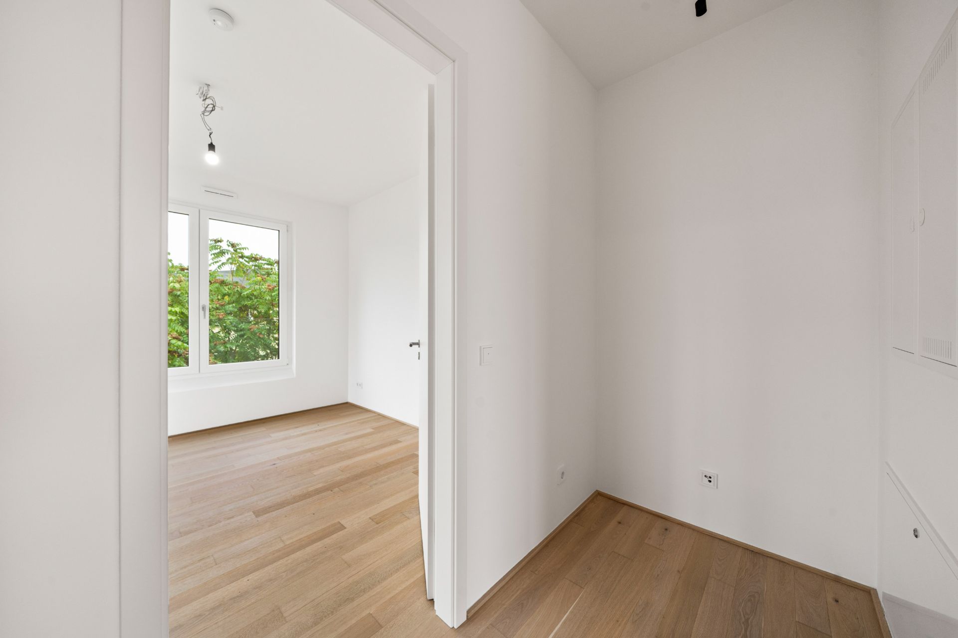 Heller Raum mit Holzboden, weiß gestrichenen Wänden und großem Fenster mit Blick ins Grüne.