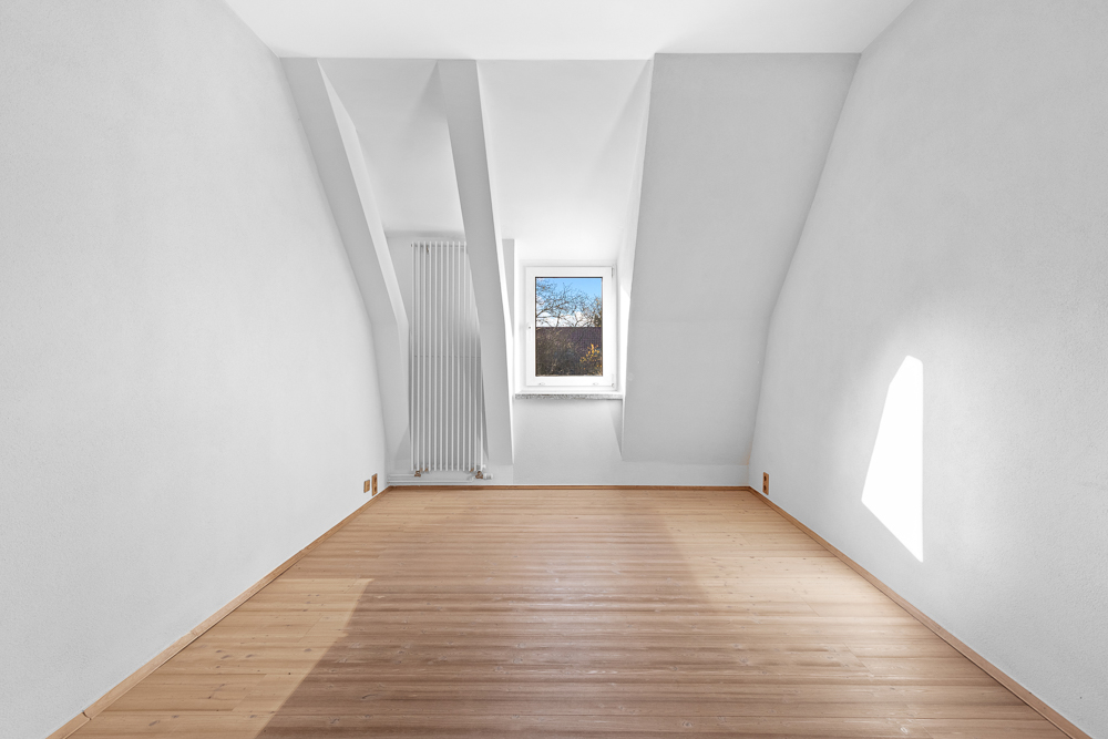 Leerer, heller Raum mit schrägen Wänden, Holzboden und einem Fenster an der Stirnseite.