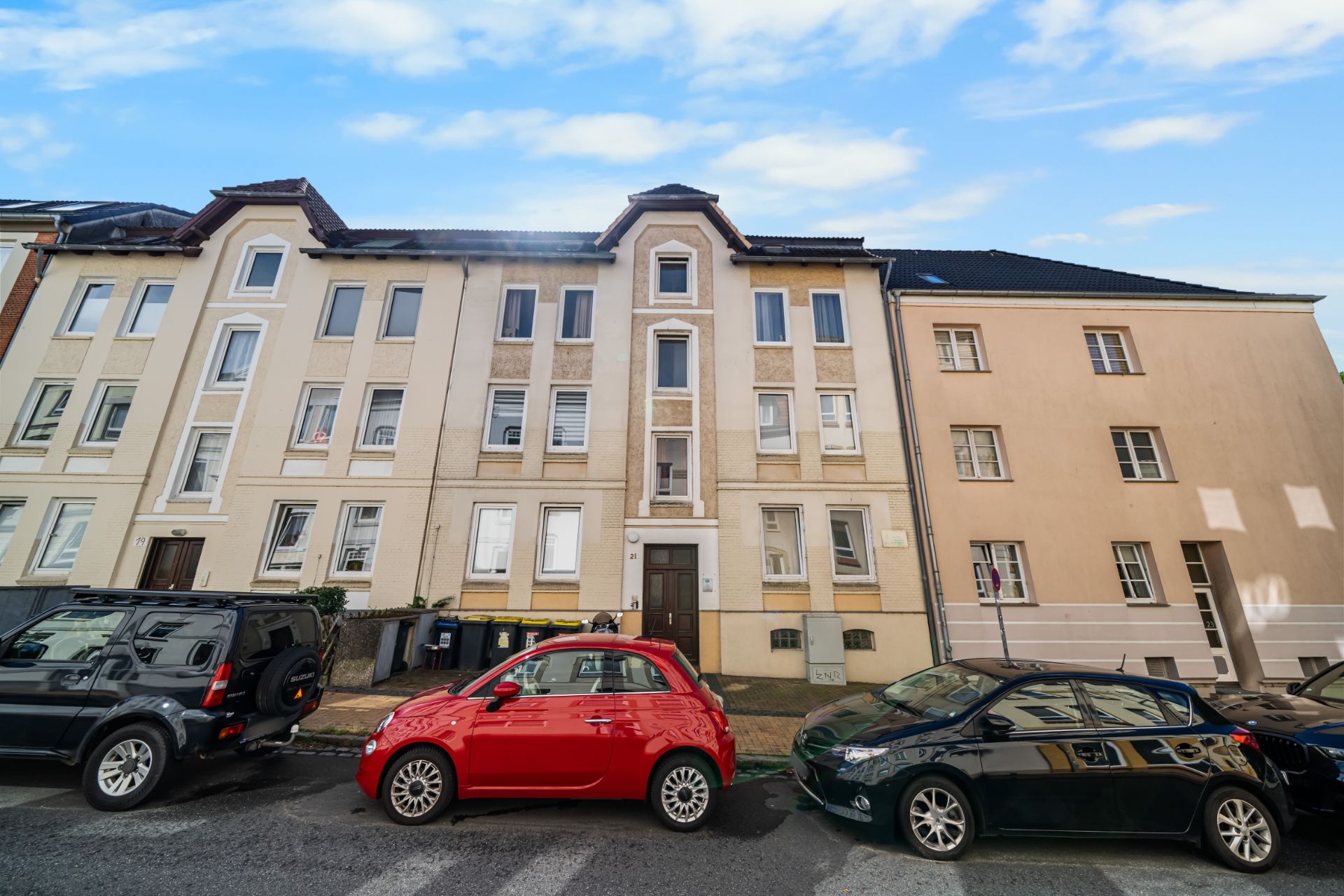 Mehrfamilienhaus mit mehreren Fenstern, drei Autos parken davor auf der Straße.
