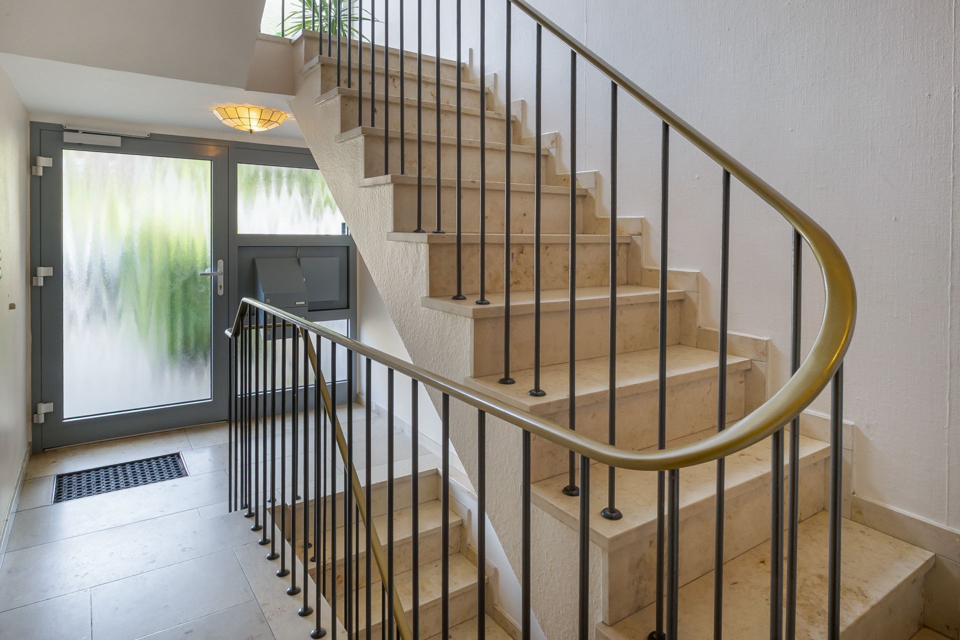 Helles Treppenhaus mit Steintreppe, Metallgeländer und Glastür mit Briefkästen.