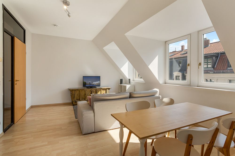 Helles Wohnzimmer mit Sofa, Esstisch, Stühlen und Fernseher, große Fenster mit Blick nach draußen.