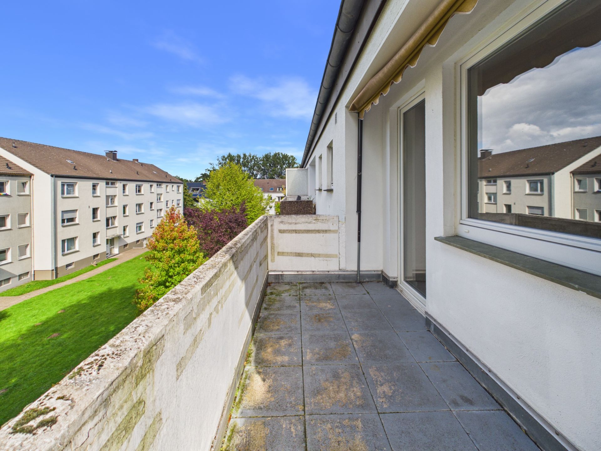 Balkon mit Fliesenboden an einem Wohnhaus, Blick auf grüne Wiese und Nachbargebäude.