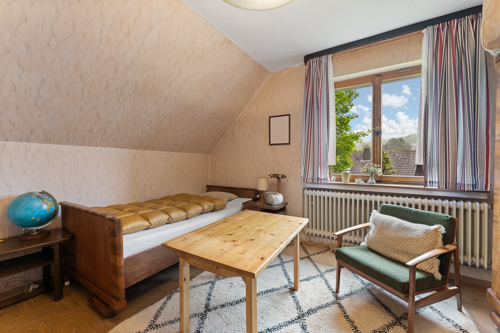 Helles Zimmer mit Einzelbett, Tisch, Sessel und großem Fenster mit Vorhängen.