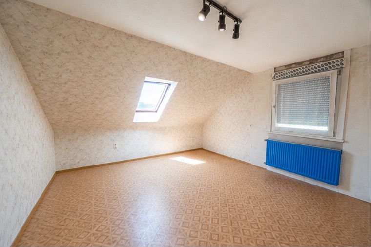 Leeres Zimmer mit Dachfenster, großem Fenster und blauem Heizkörper unter der Schräge.
