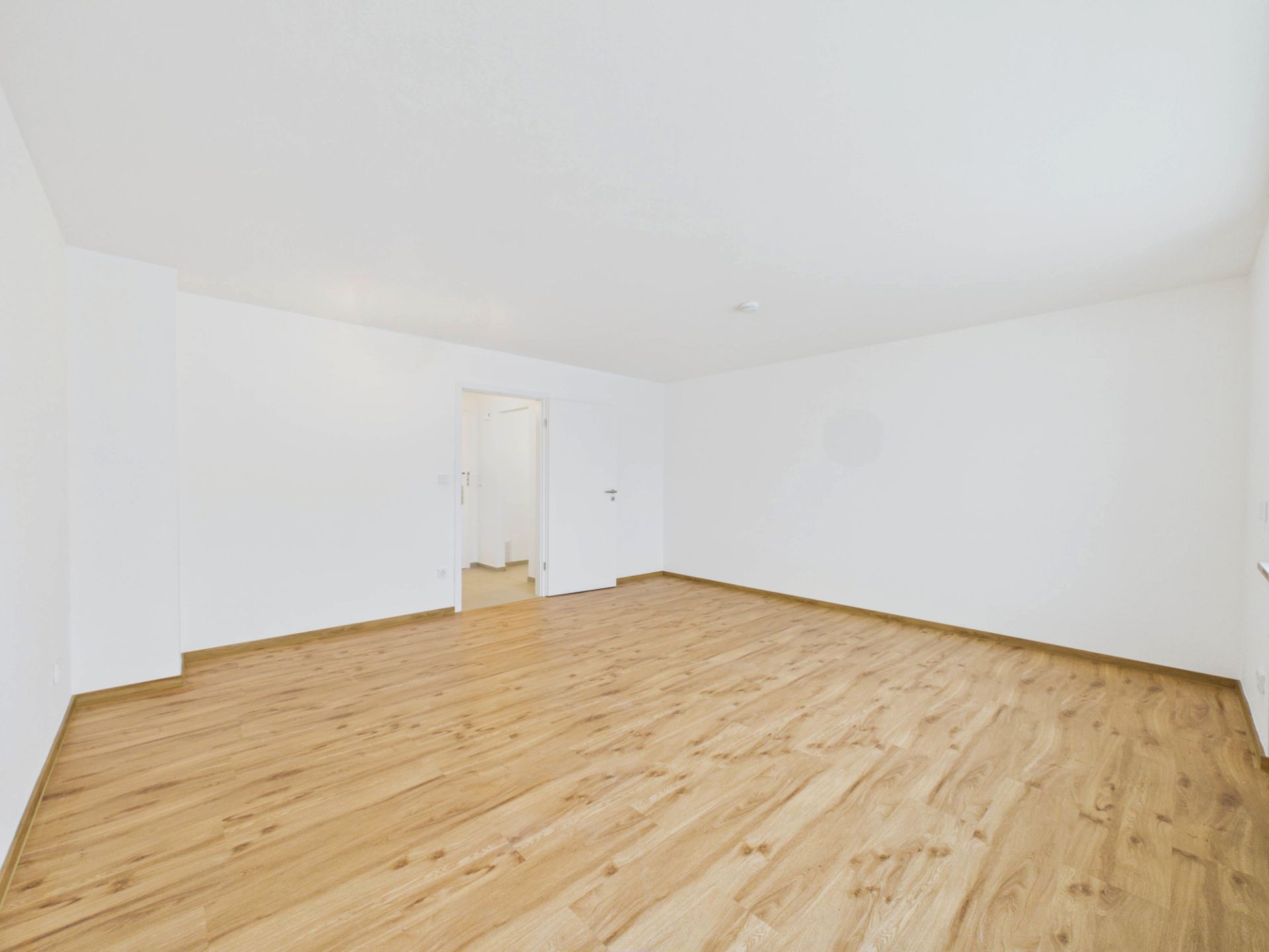 Heller, leerer Raum mit Holzfußboden und weißen Wänden, Tür im Hintergrund.