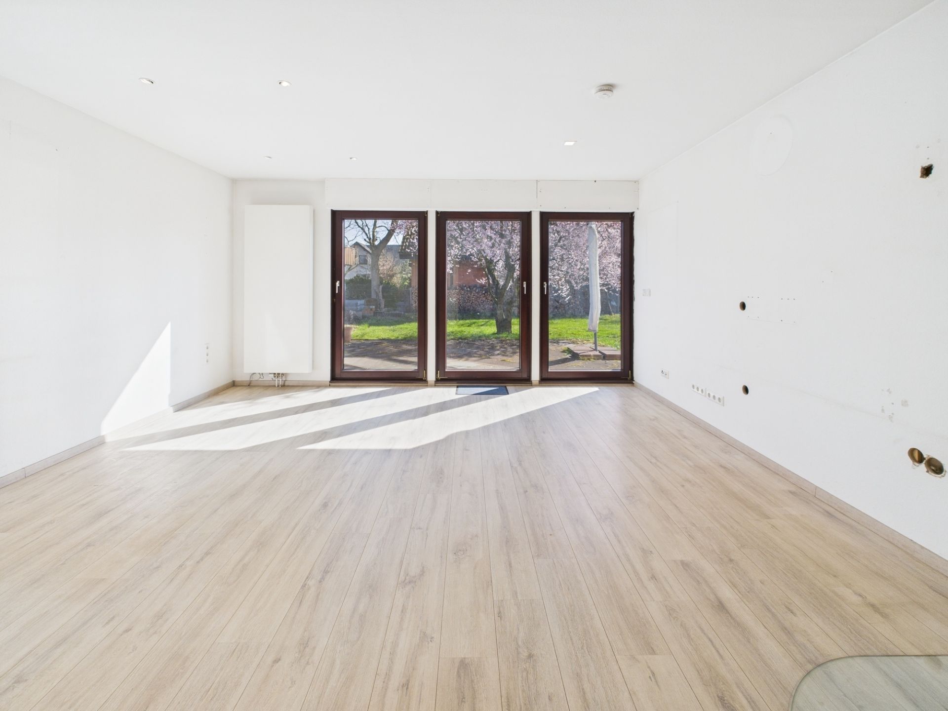 Leerer, heller Raum mit Holzfußboden und großen Fenstern zum Garten.