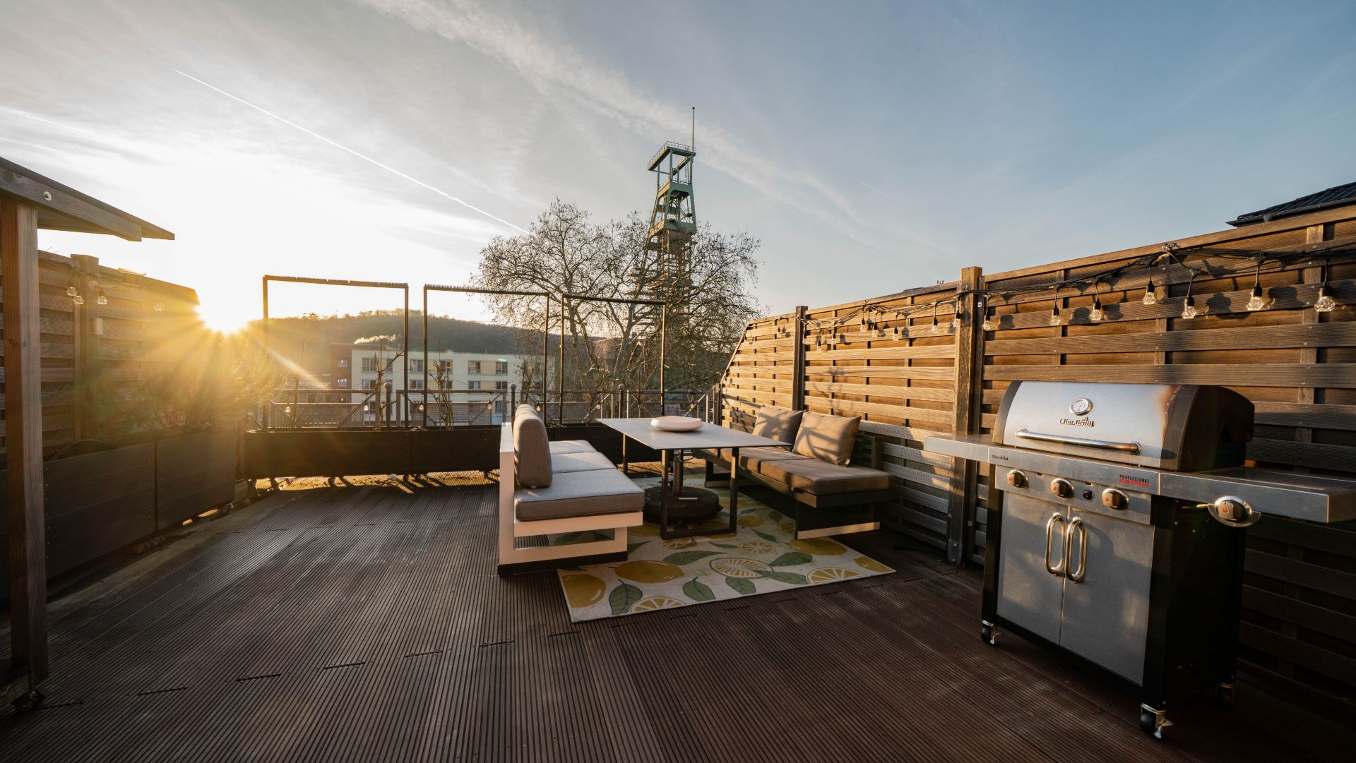 Dachterrasse mit Sitzecke, Teppich, Grill und Holzwänden bei Sonnenuntergang.