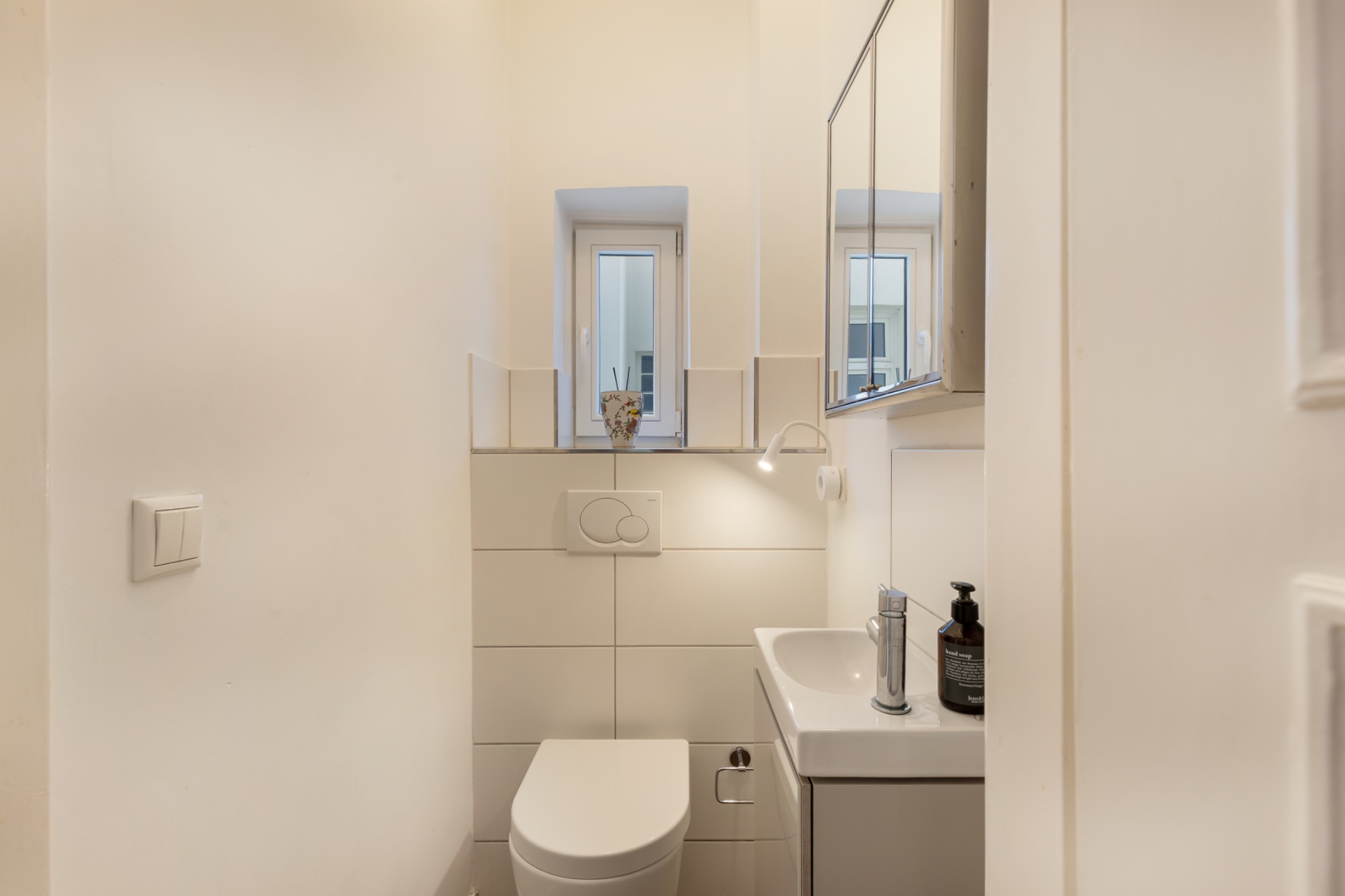 Kleines, helles Badezimmer mit WC, Waschbecken, Spiegel und Fenster.
