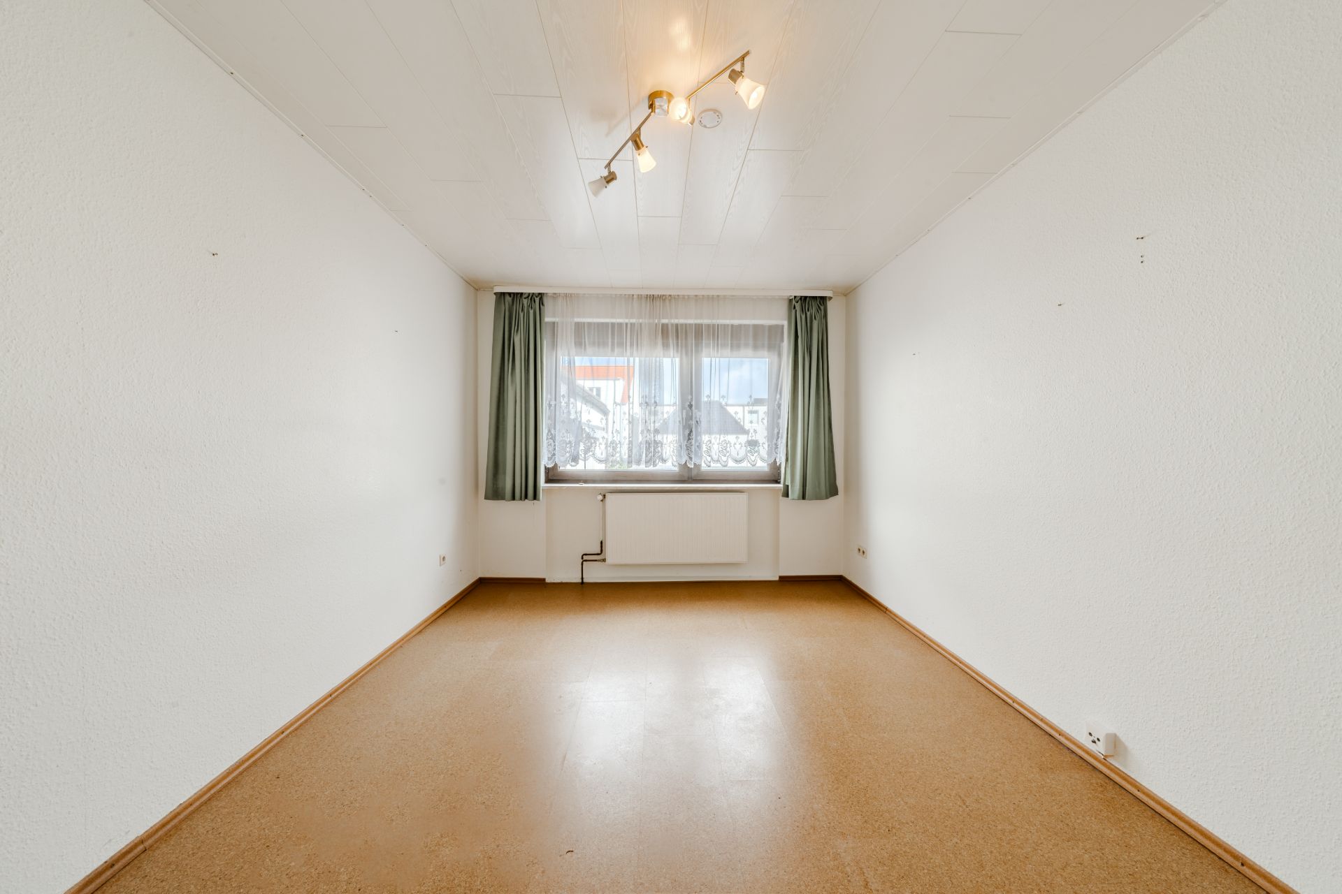 Leerer rechteckiger Raum mit Fenster, Vorhängen, Deckenleuchte und hellem Boden.