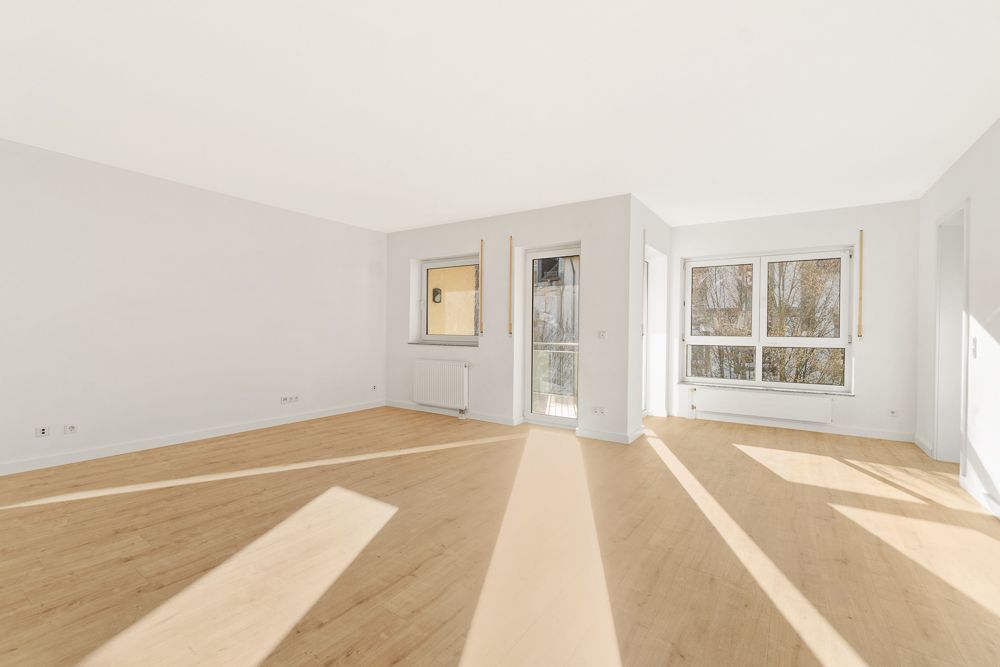Heller, leerer Raum mit großen Fenstern, weißer Wand und hellem Holzboden.