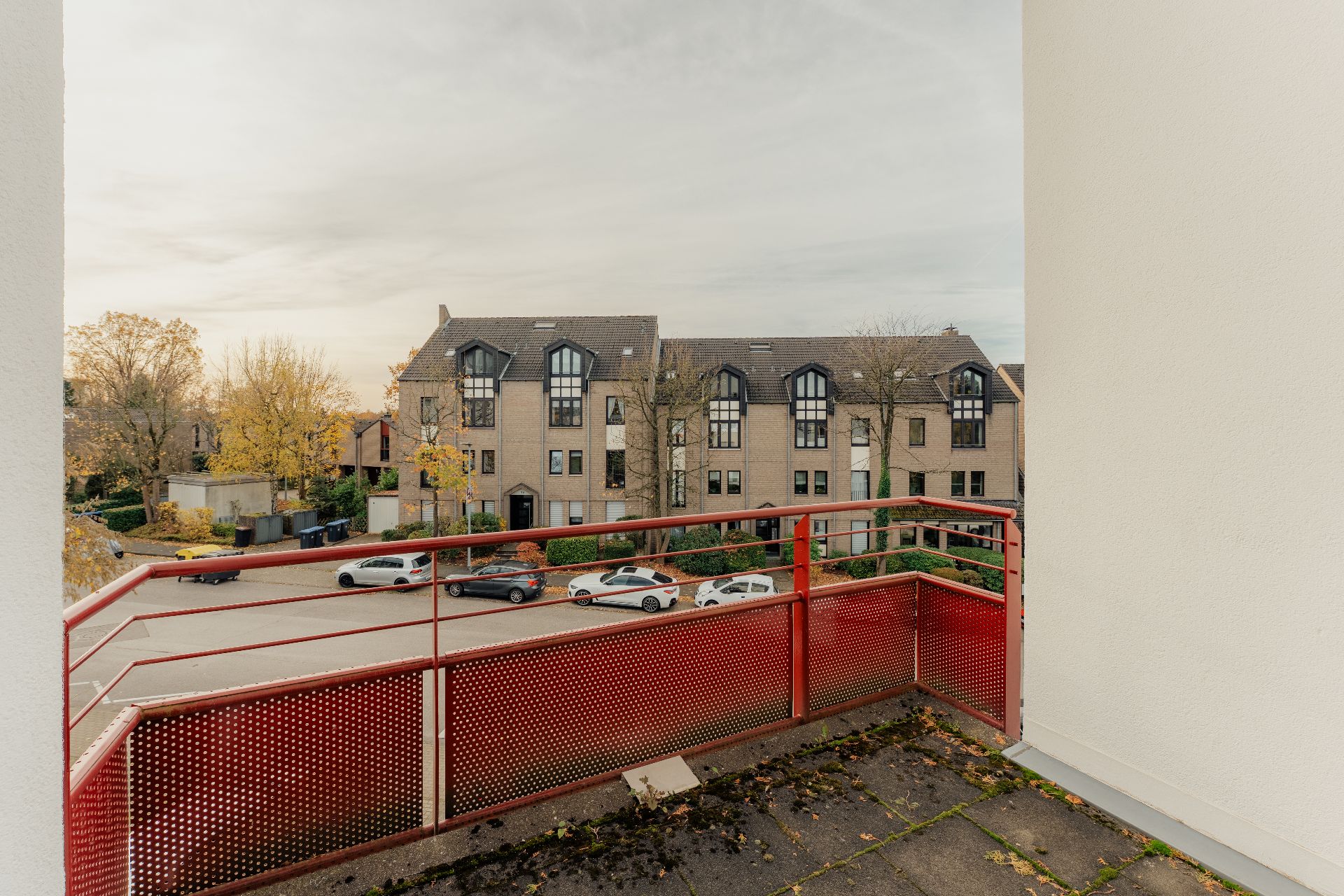 Blick von einem Balkon mit rotem Geländer auf eine ruhige Wohnstraße und mehrere geparkte Autos.