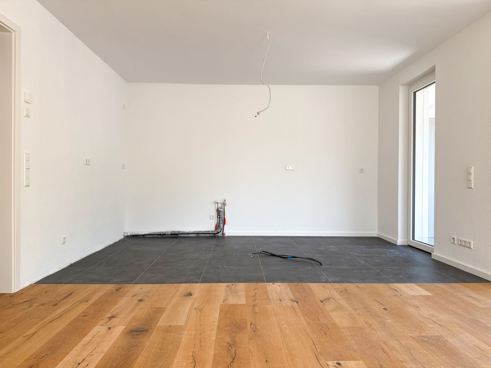 Leerer Raum mit Parkett- und Fliesenboden, weiße Wände und große Fenster, sichtbare Kabel am Boden.
