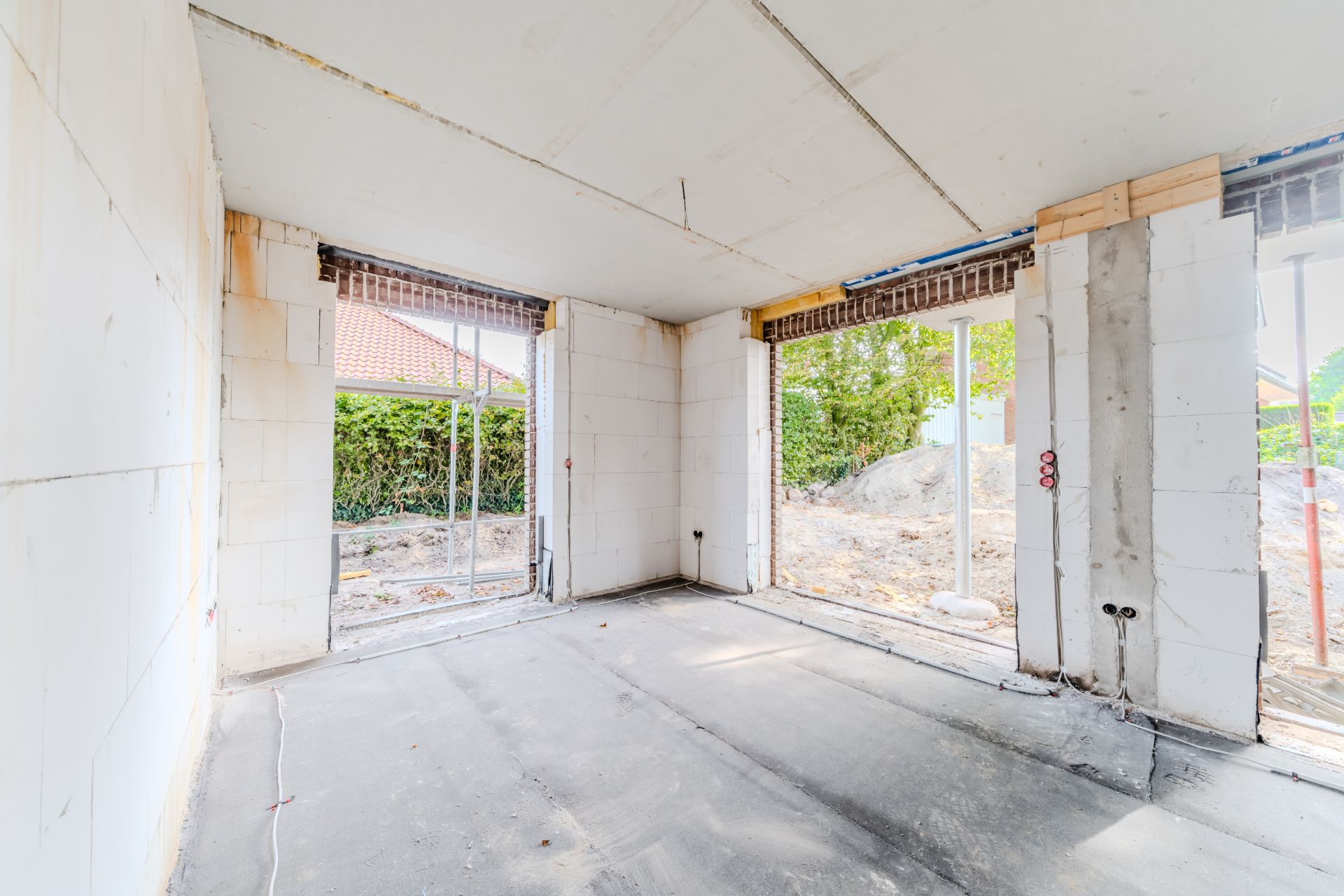 Heller, unfertiger Raum mit großen Fensteröffnungen und Blick auf Garten und Baustelle.