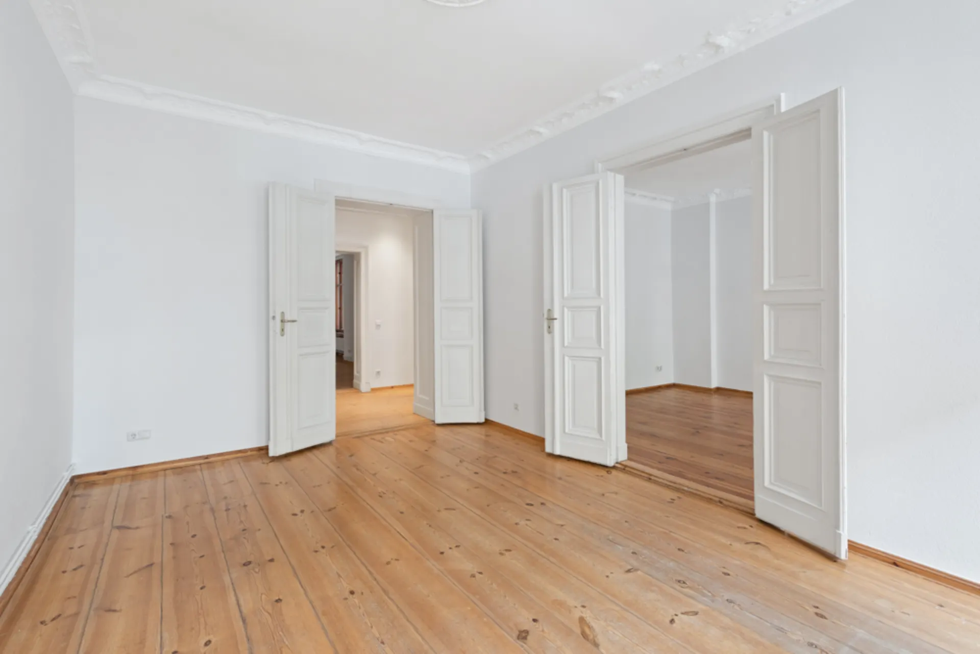 Leerer Raum mit Holzboden, weißen Wänden und offenen Doppeltüren.
