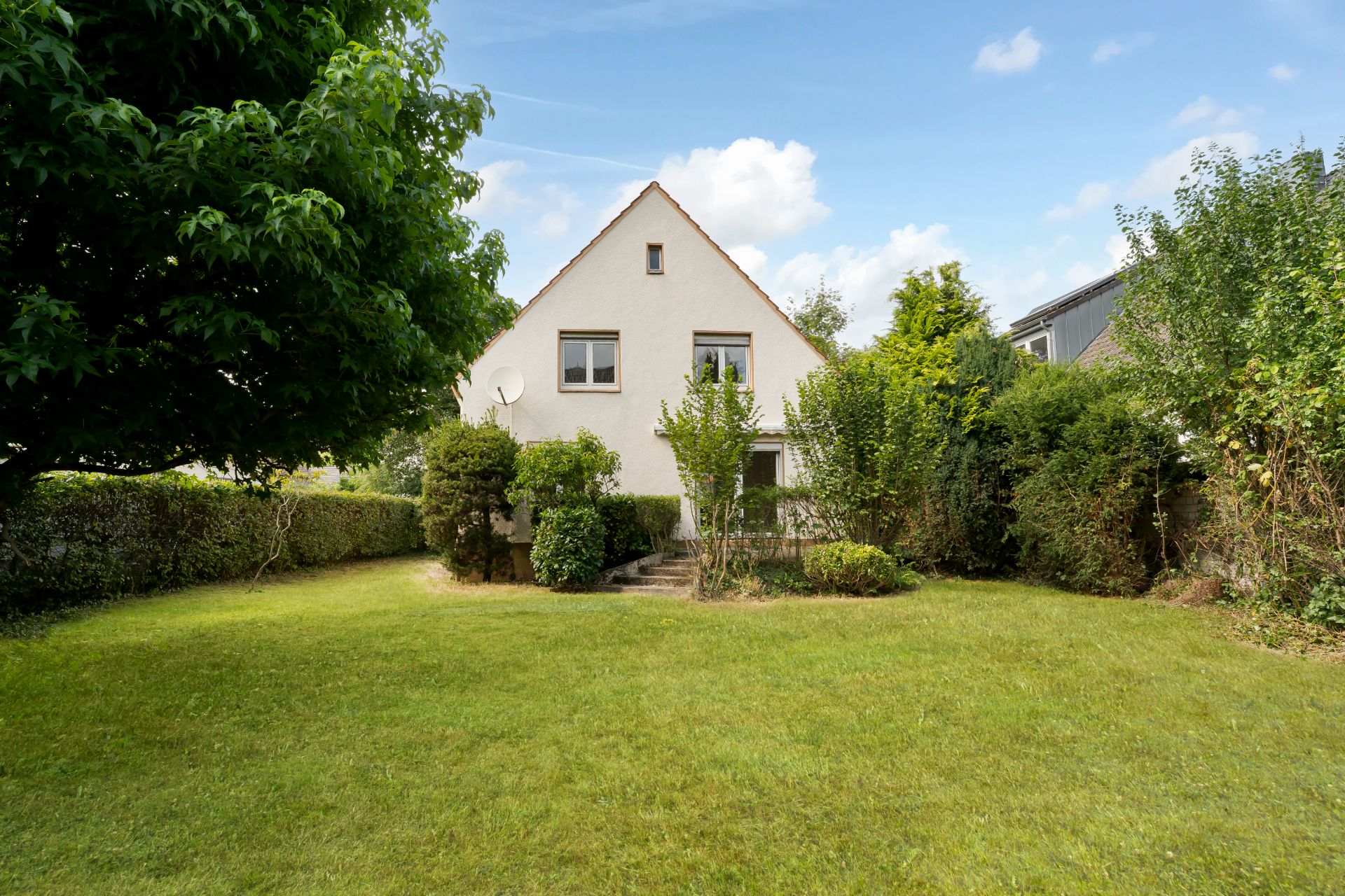 Einfamilienhaus mit Garten, Bäumen und Rasenfläche an sonnigem Tag.