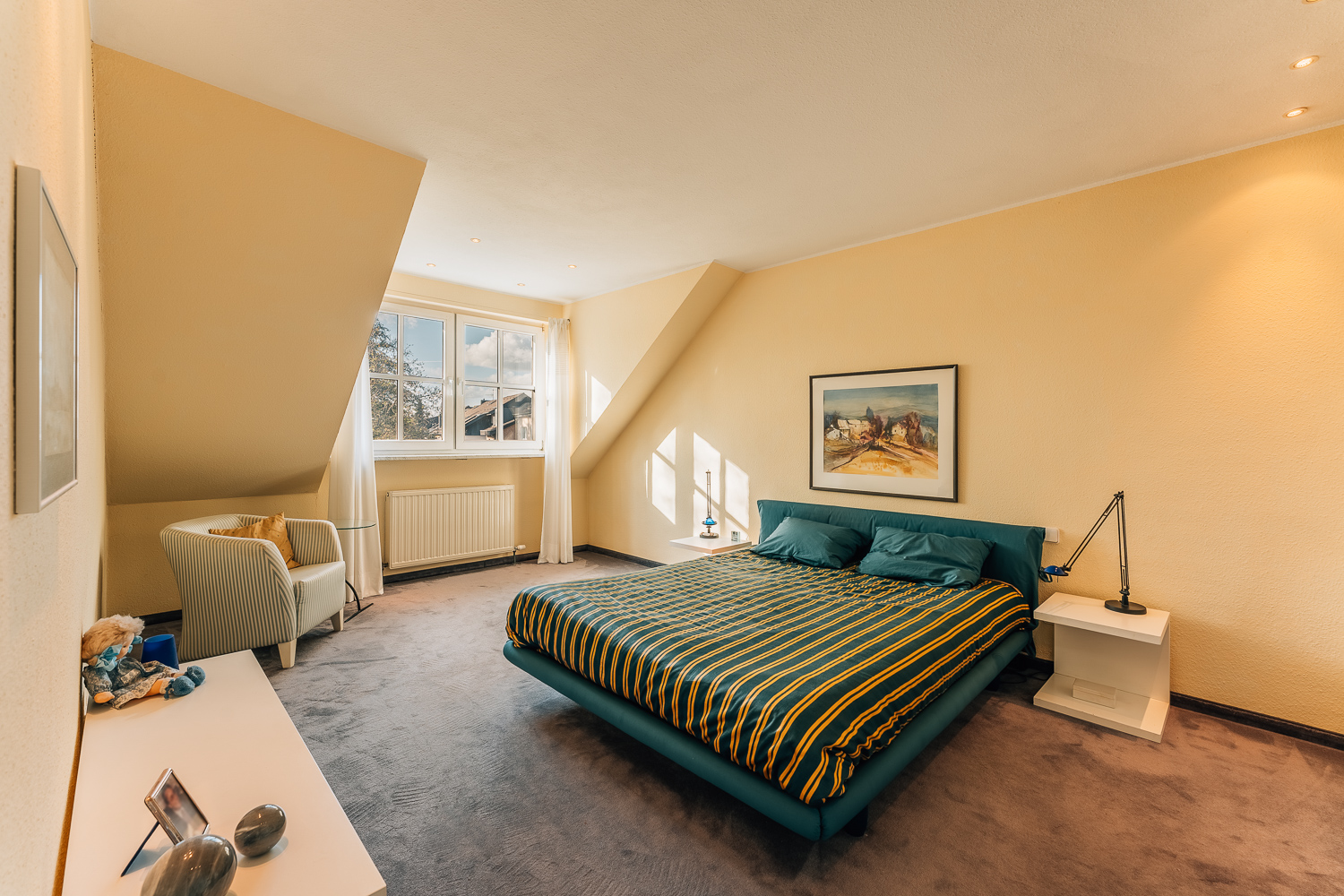 Helles Schlafzimmer mit Doppelbett, Sessel, Bild an gelber Wand und großem Fenster.
