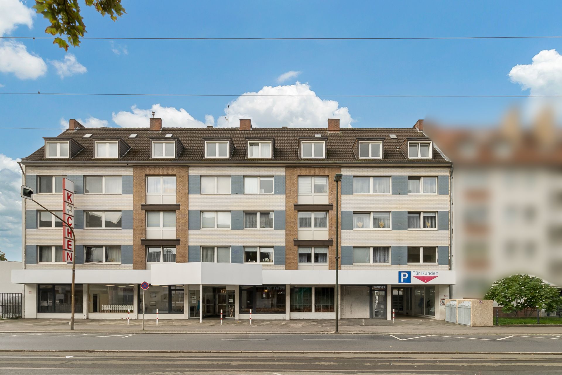 Vierstöckiges Wohnhaus mit mehreren Fenstern und Parkplätzen davor an einer Straße.