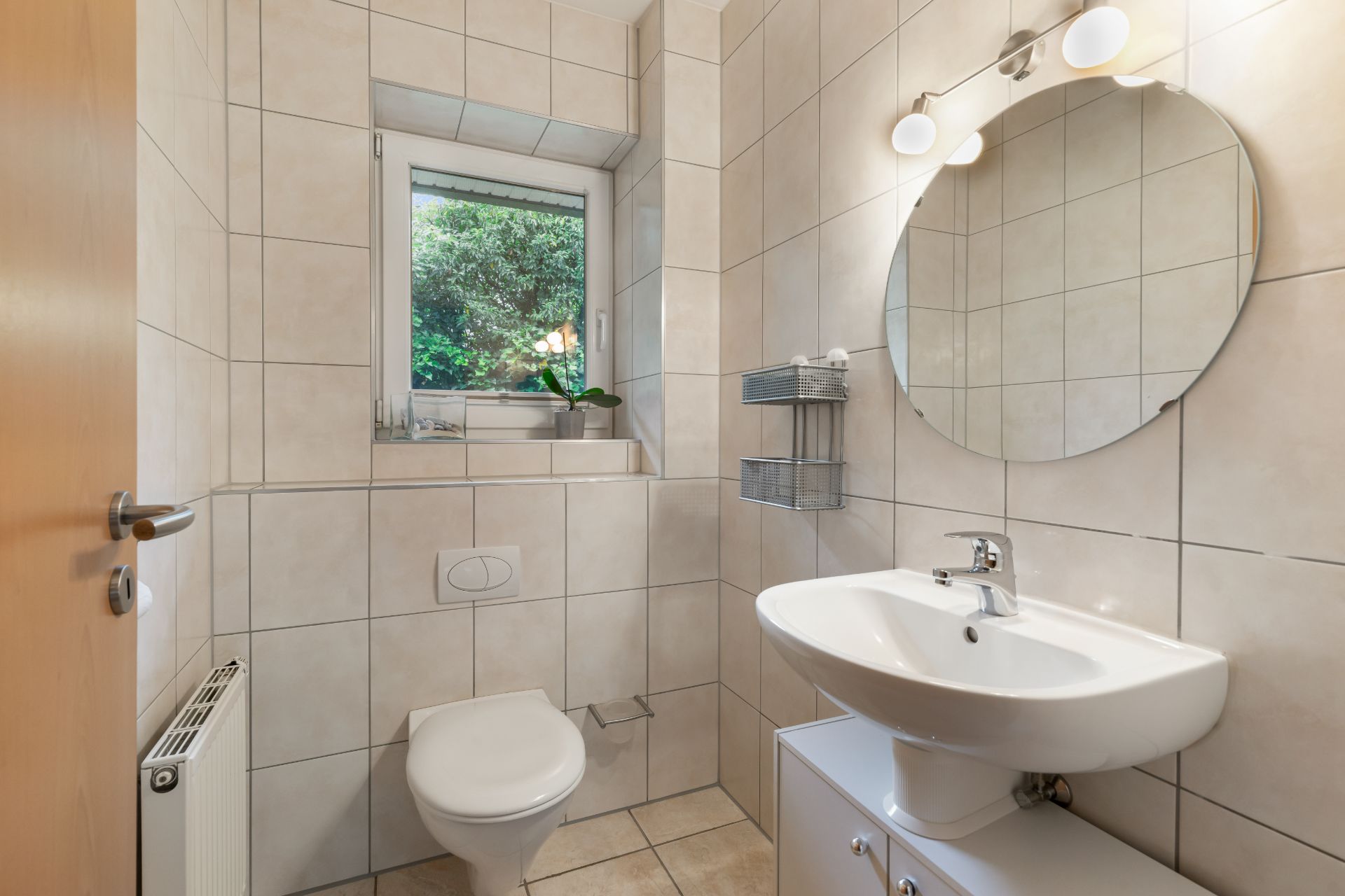 Helles Badezimmer mit WC, Waschbecken, Spiegel und Fenster mit Blick ins Grüne.