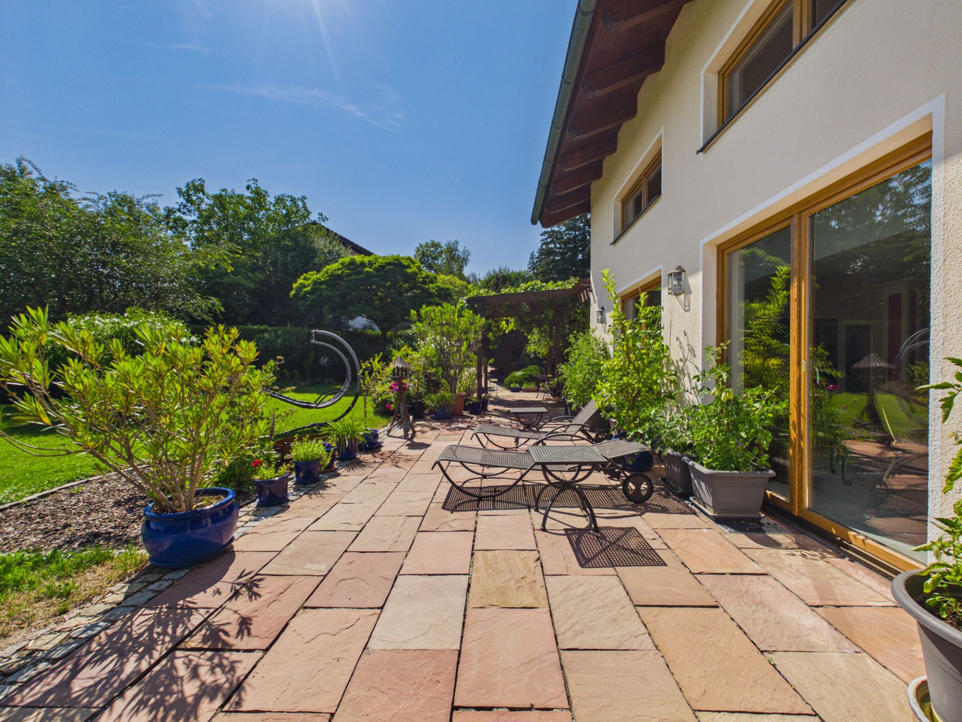 Große Terrasse mit Liegestühlen, Pflanzen in Töpfen und Blick auf einen Garten bei Sonnenschein.