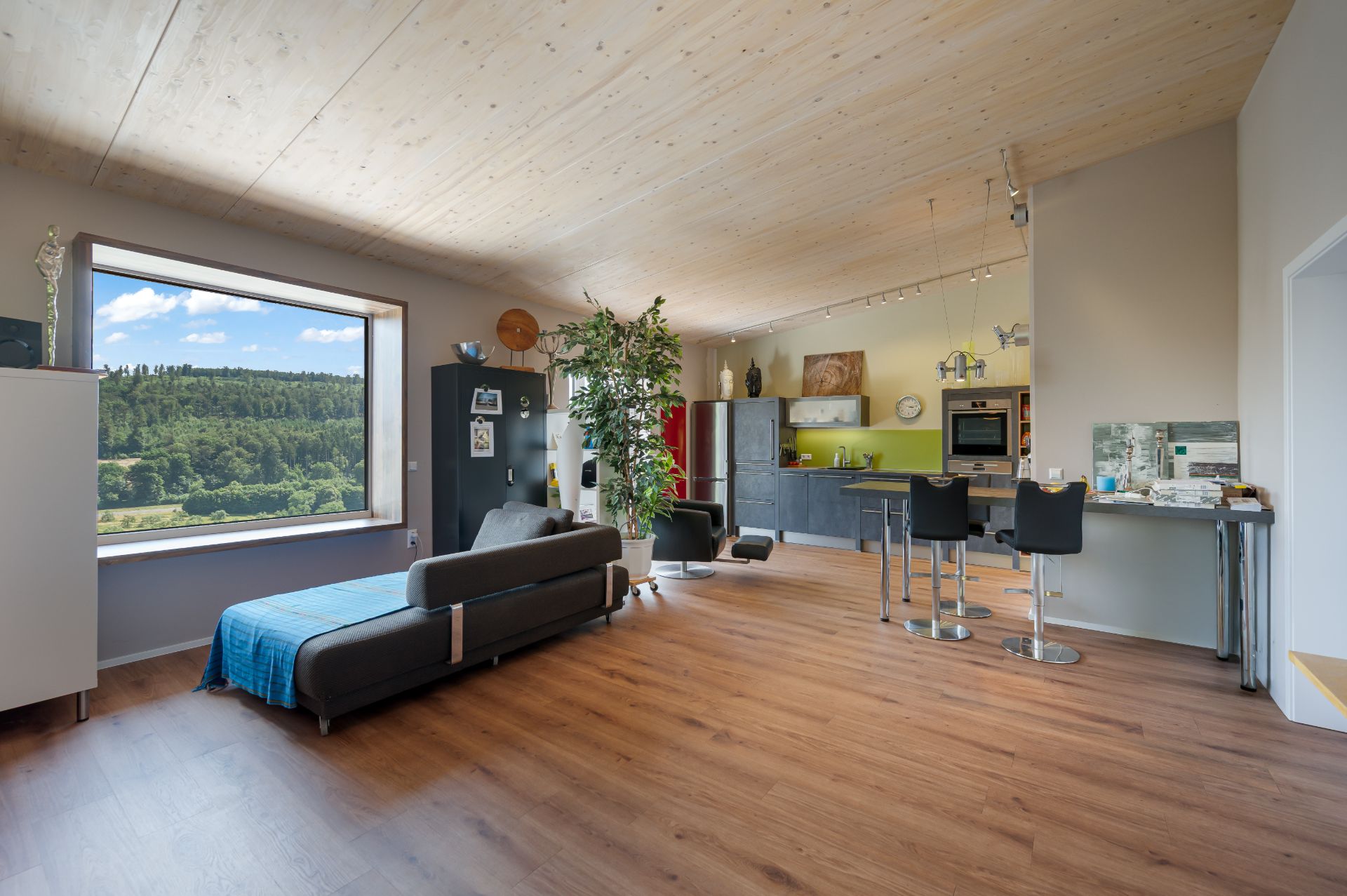 Helles Wohnzimmer mit großer Fensterfront, Couch, offener Küche und Holzboden.