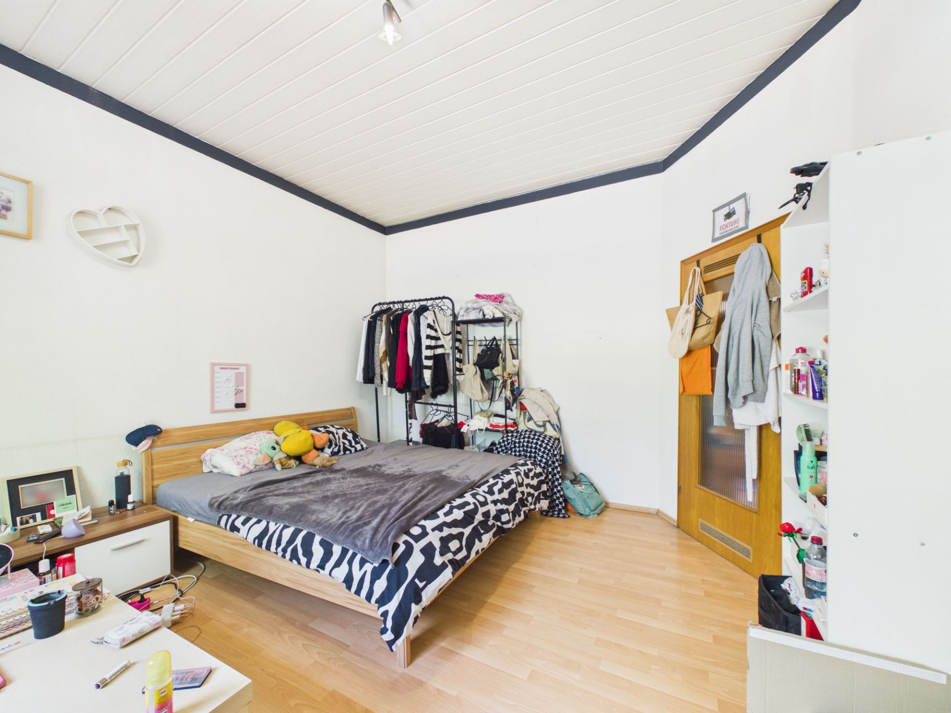 Helles Schlafzimmer mit Bett, Kleiderständer, Regal und Holztür.