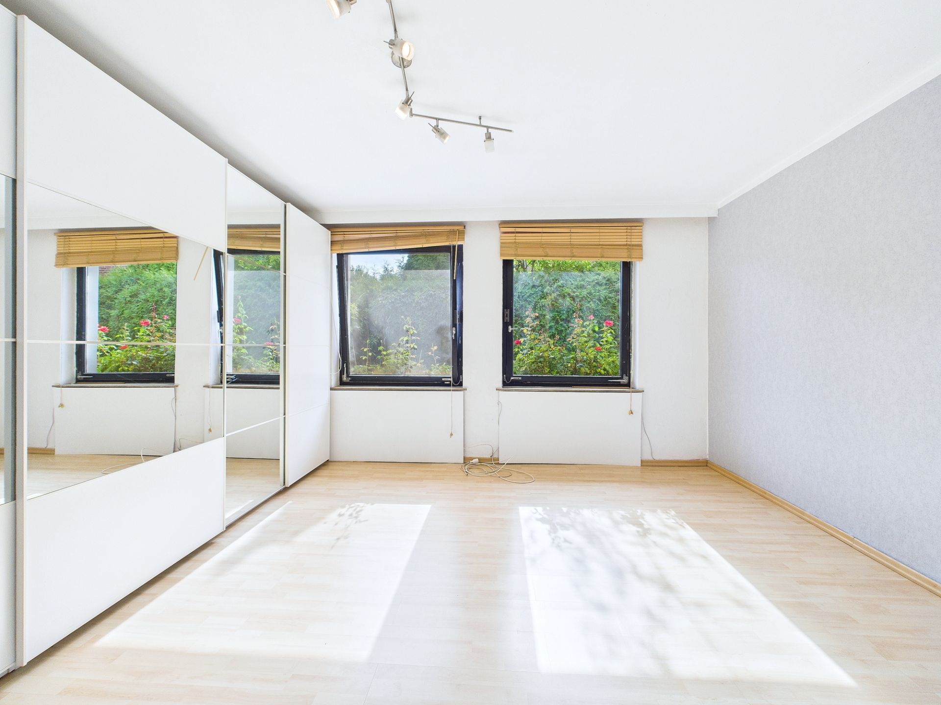 Helles Zimmer mit Parkettboden, drei Fenstern und einem großen Spiegel-Schrank an der Wand.