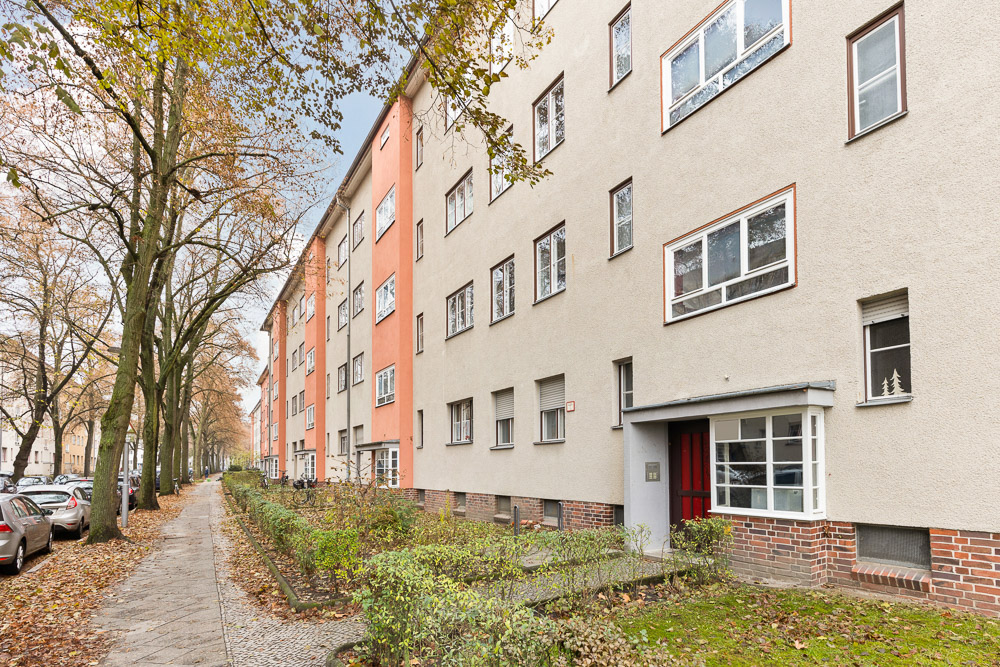 Mehrfamilienhaus mit mehreren Fenstern und einem Eingang, Gehweg und Bäumen davor.
