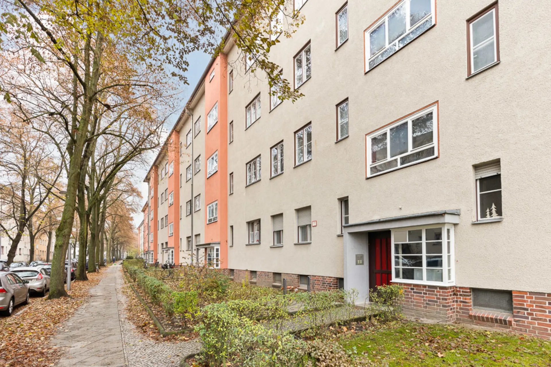 Mehrfamilienhaus mit mehreren Fenstern und einem Eingang, Gehweg und Bäumen davor.