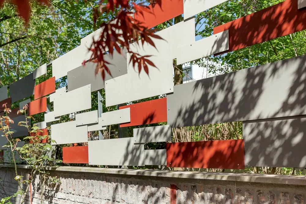 Moderner Gartenzaun aus abwechselnd roten und grauen Paneelen mit Bäumen im Hintergrund.