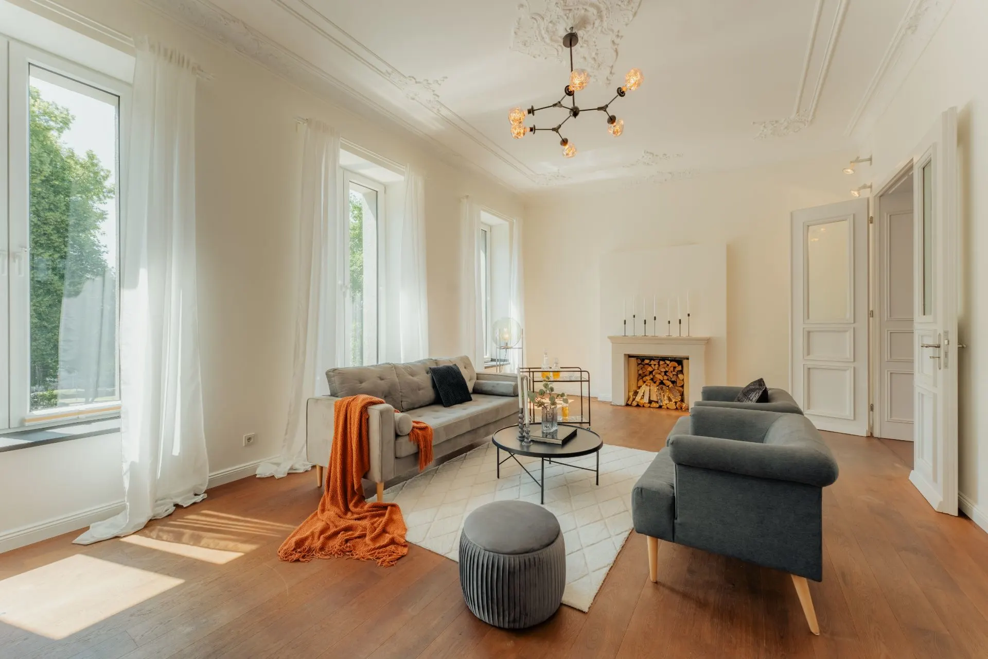 Helles Wohnzimmer mit grauem Sofa, Kamin, Holzboden und großen Fenstern mit weißen Vorhängen.