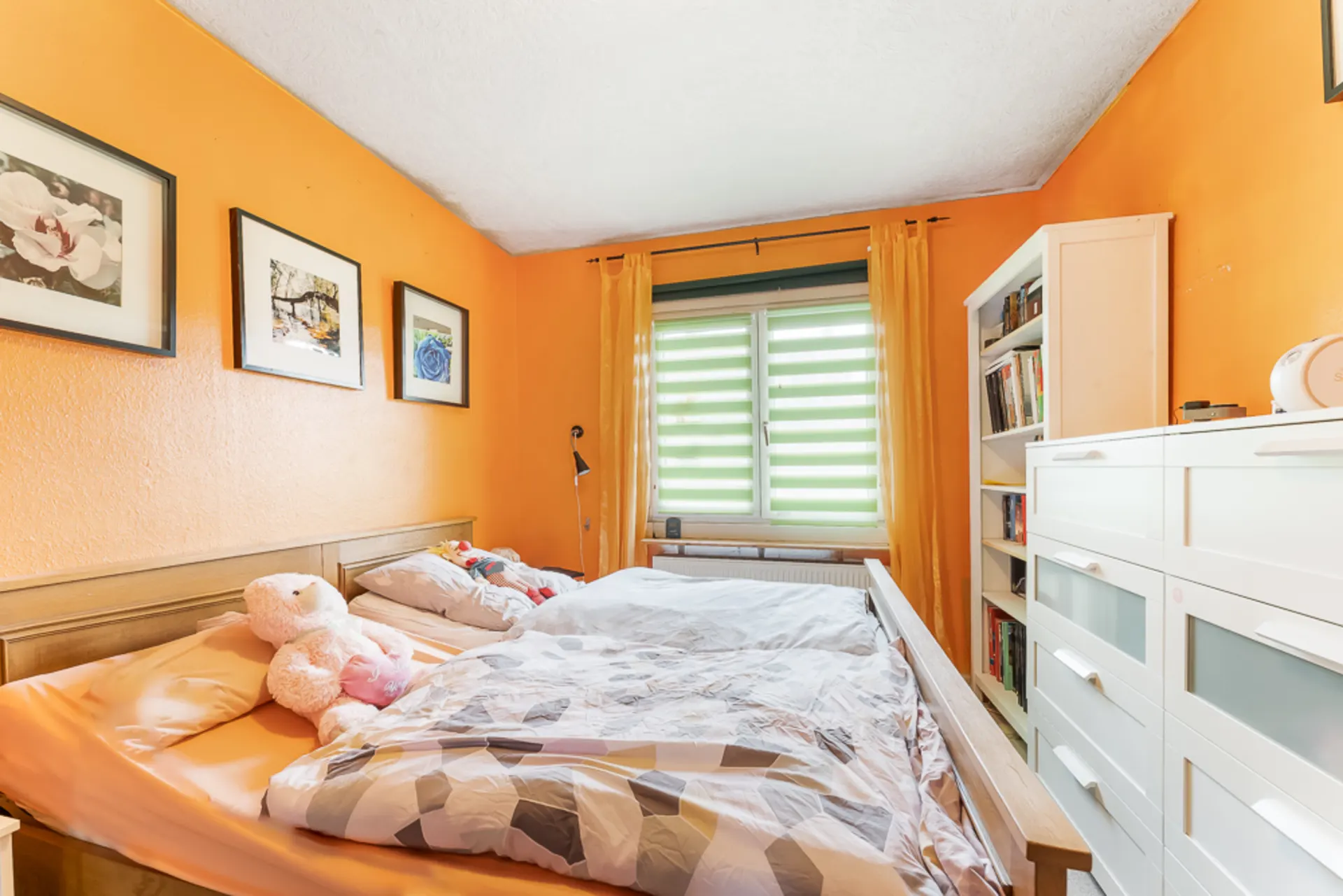 Helles Schlafzimmer mit Doppelbett, orangenen Wänden, Fenster, Kommode und Bücherregal.