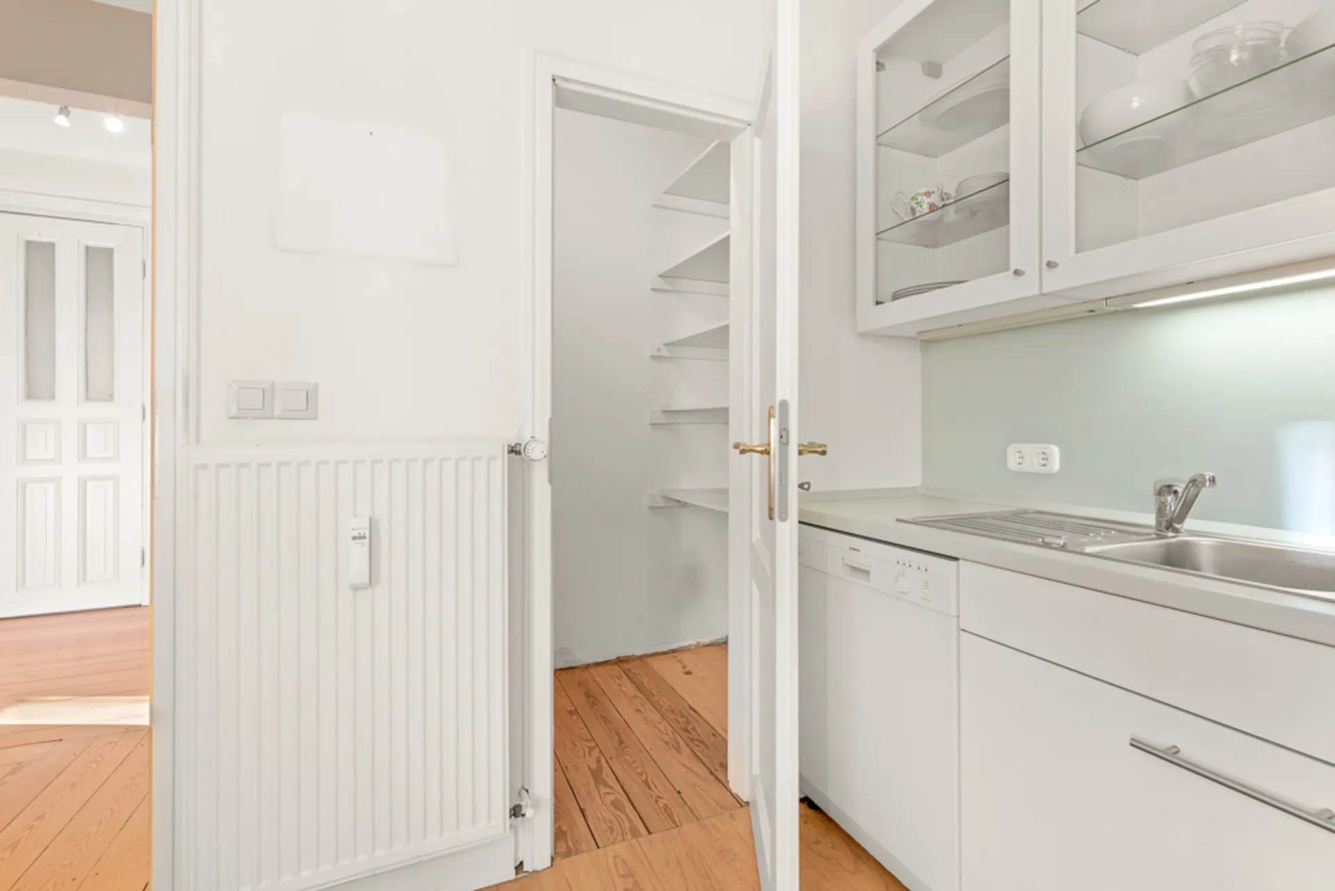 Helle Küche mit Spüle, Geschirrspüler und Glastüren, nebenan eine Speisekammer mit Regalen.